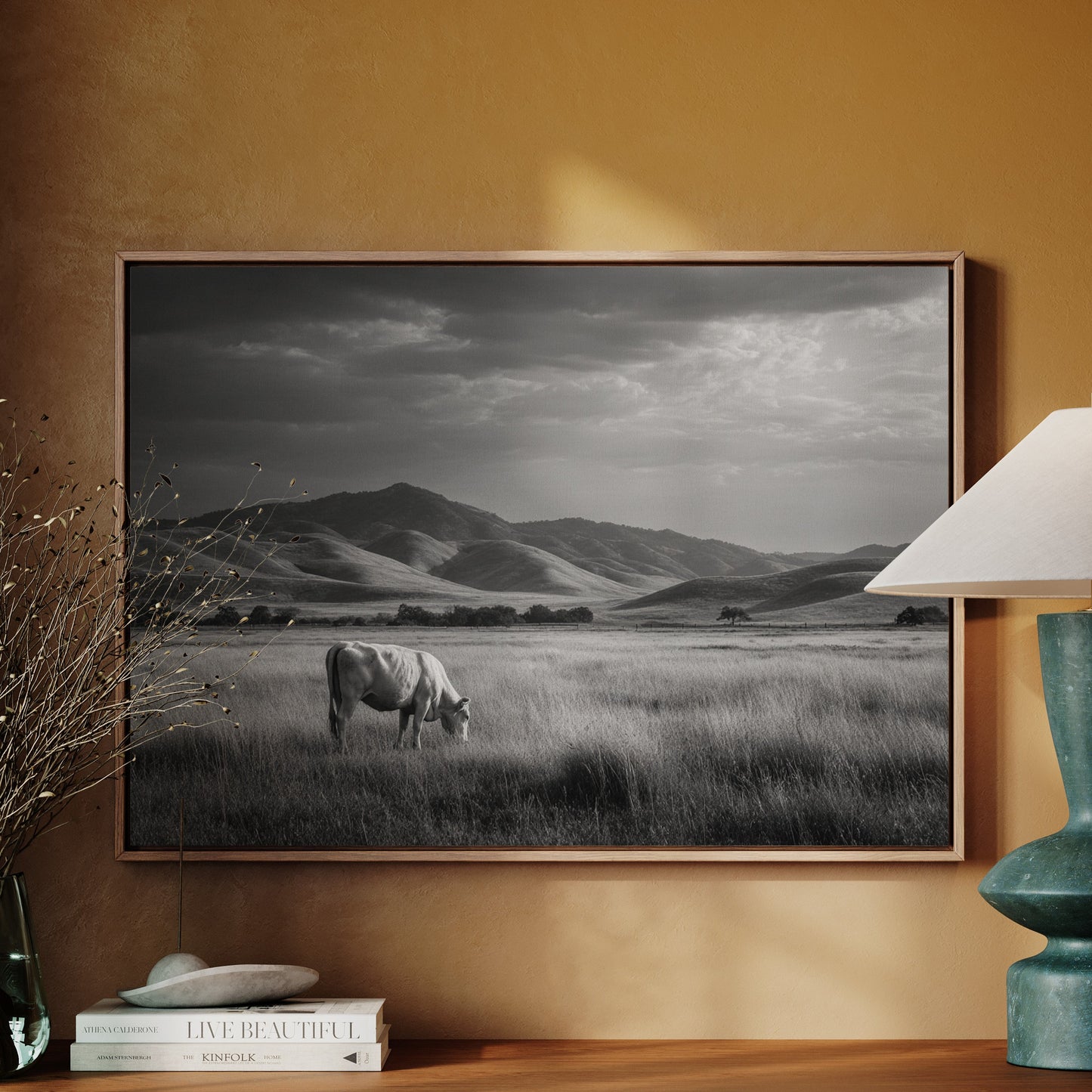 Framed black and white landscape of a cow in a field on a wall above a wooden cabinet with decor items.