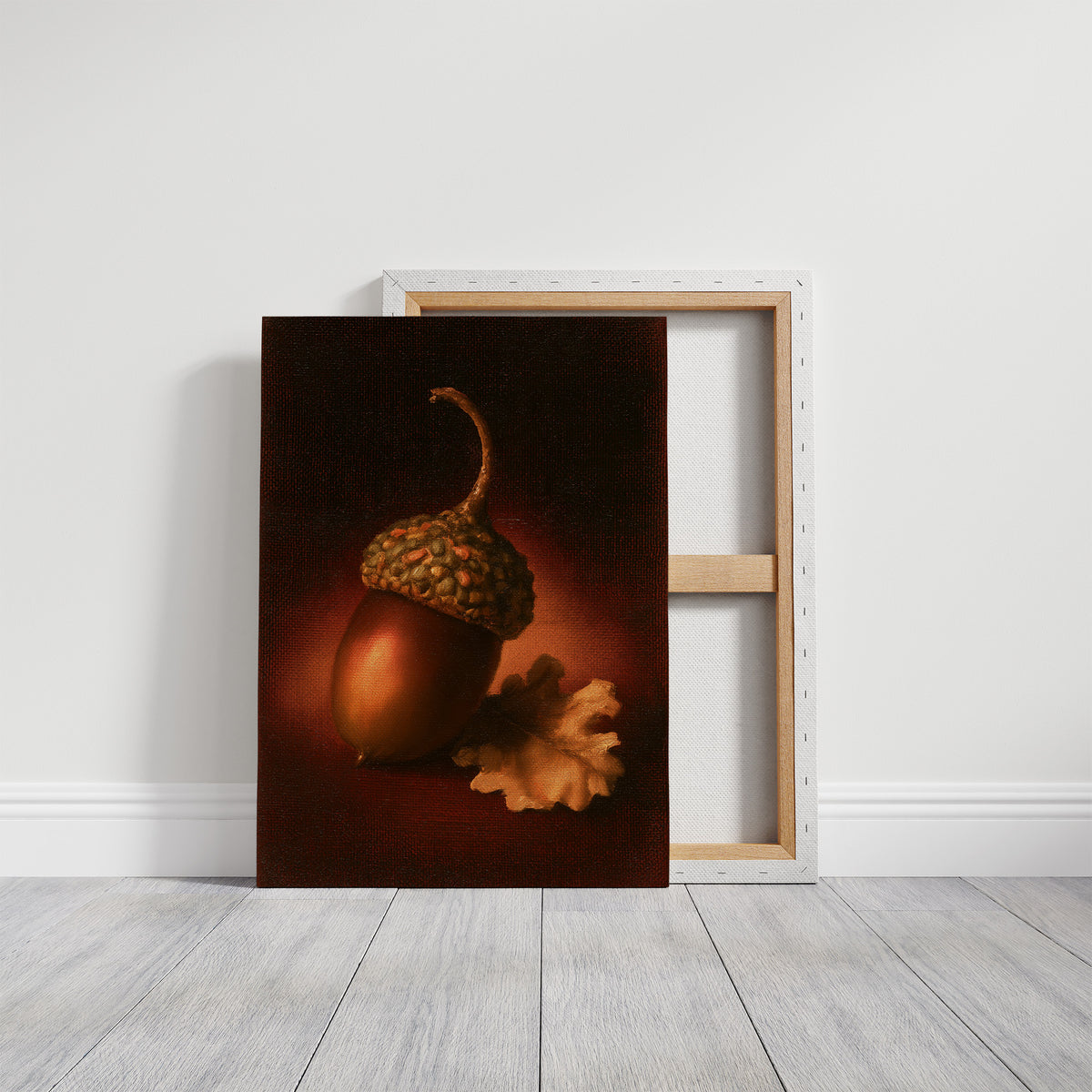 A Single Acorn in Candlelight, one acorn resting beside a curled oak leaf on a dark brown ground, lit from one side and painted in chestnut, amber and deep shadow, on a gallery canvas with wrapped edges leaning against a white wall