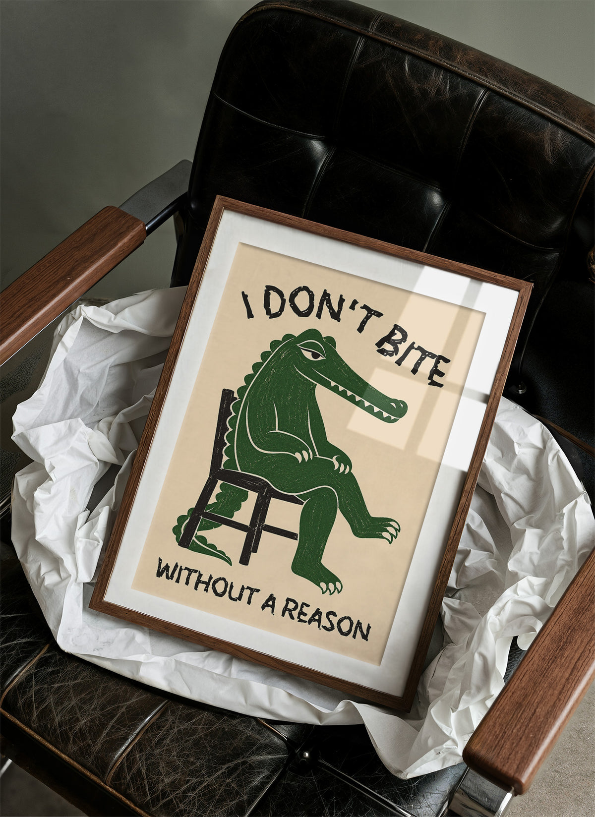 Bites Only With a Reason, a crocodile sitting upright in a plain wooden chair, in deep green on a cream ground, with a bold lettered line above reading I DON'T BITE and one below reading WITHOUT A REASON, in a wood frame on a black leather armchair beside crumpled white tissue