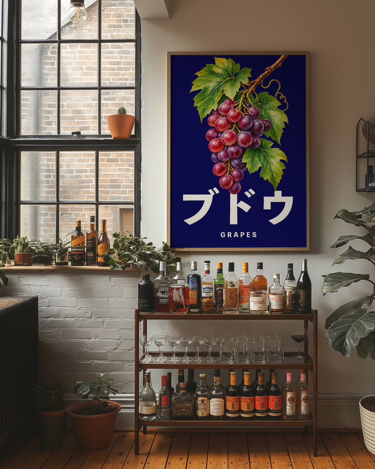 Clusters Heavy Over Midnight Water, a cluster of rose and purple grapes with green leaves hanging from a stem on a deep navy ground, with the Japanese katakana set in large white letters and the word GRAPES in small capitals beneath, in a wood frame on a brick wall above a bar cart of bottles and plants