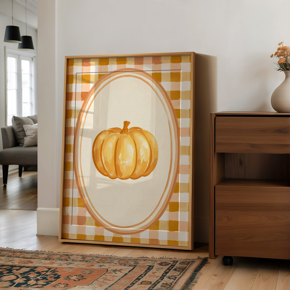 Dressed Up for a Quiet Afternoon, a single orange pumpkin in a cream oval medallion framed by a yellow and rust gingham border, painted in mustard, cream and burnt orange, in a wood frame on a wall beside a walnut cabinet and an open doorway