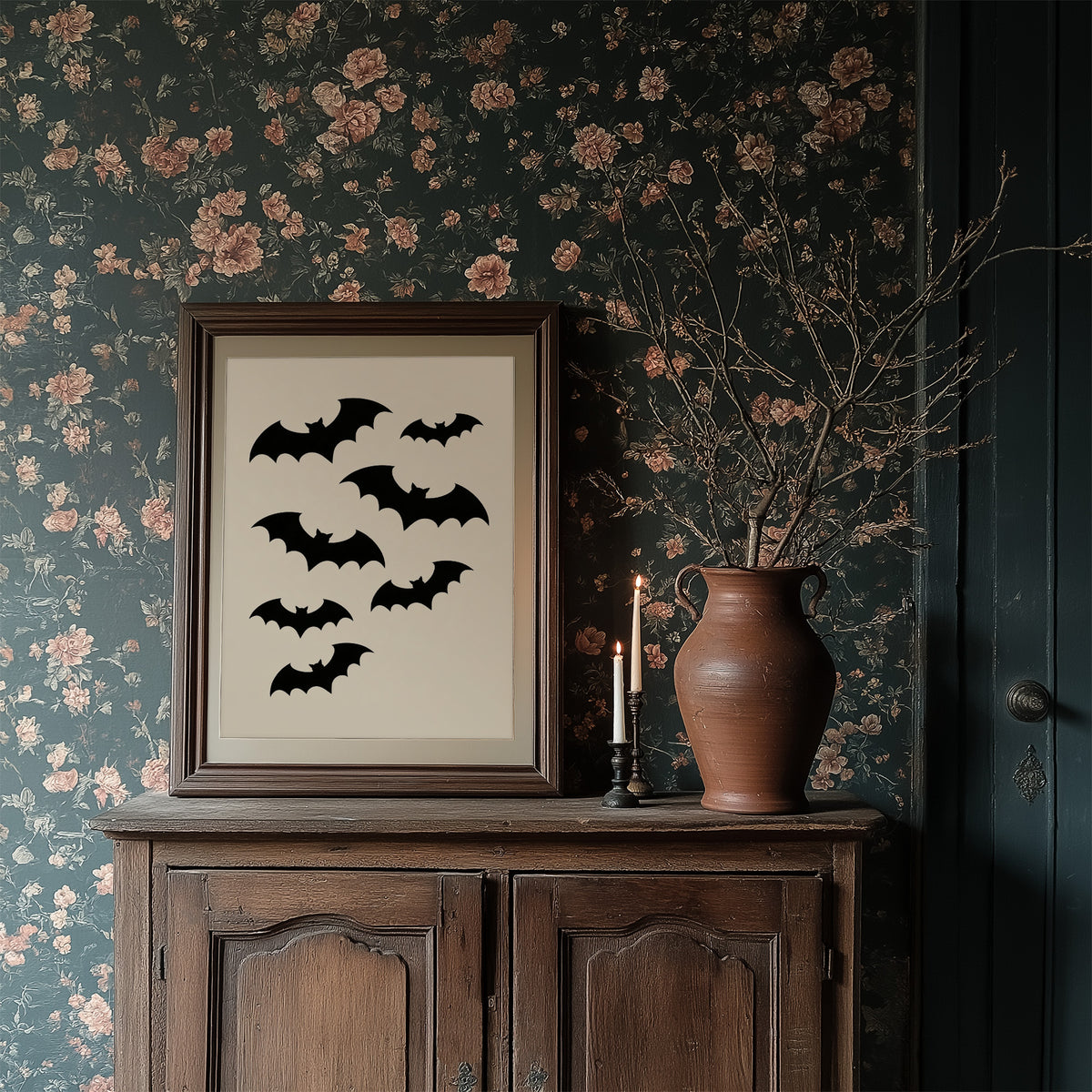 Evening Flight in Silhouette, a loose flock of black bat silhouettes spread across a cream ground in a simple, flat graphic arrangement, in a dark wood frame on a floral wallpaper wall above a rustic cabinet and a vase