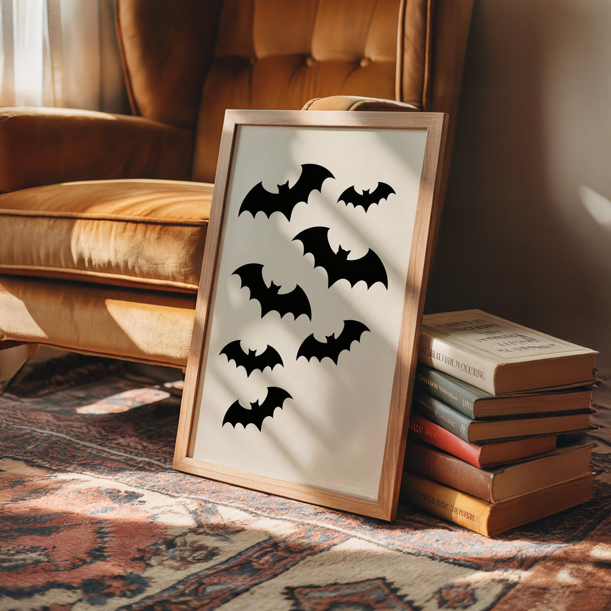 Evening Flight in Silhouette, a loose flock of black bat silhouettes spread across a cream ground in a simple, flat graphic arrangement, in a wood frame on a leather armchair beside a stack of books