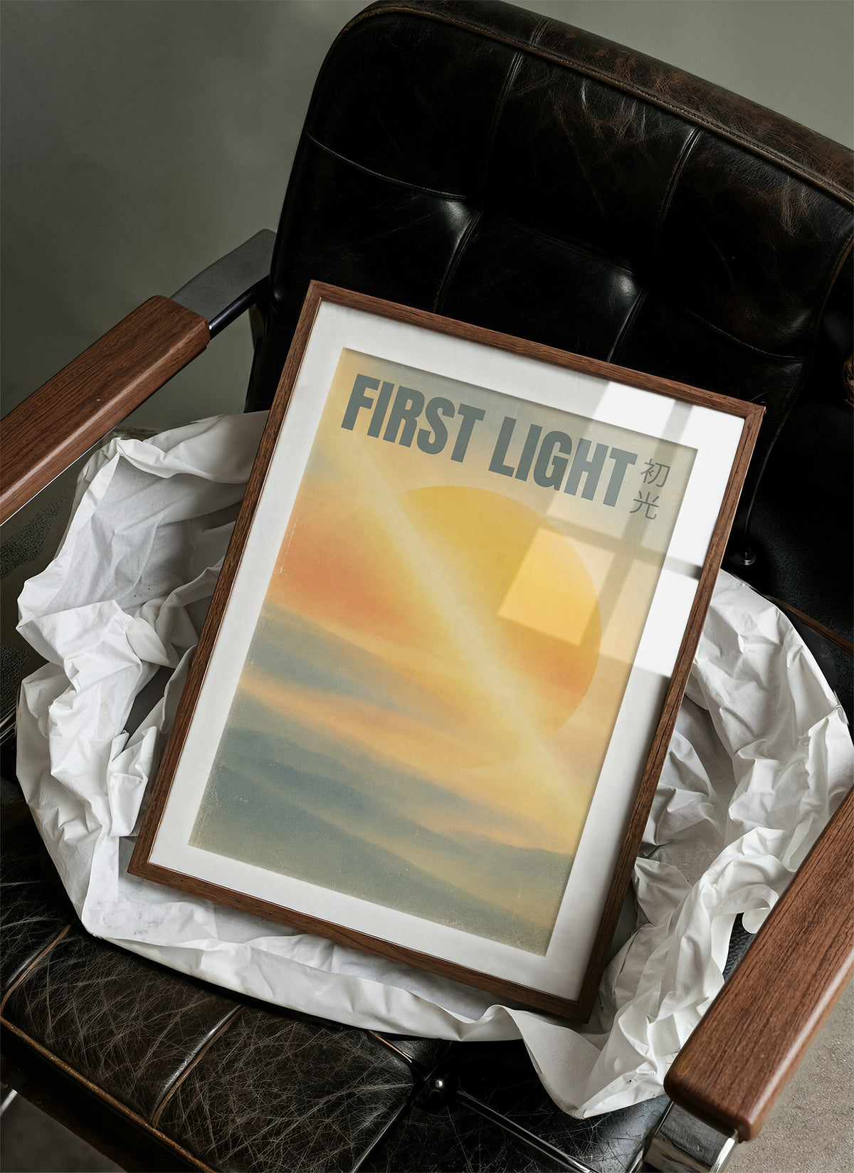 First Light Over a Quiet Sea, a pale sunrise over a soft sea, a warm yellow sun glowing above a band of blue water and peach clouds, with the words FIRST LIGHT in gray capitals across the top and a small Japanese word beside them, in a wood frame on a black leather armchair beside crumpled white tissue