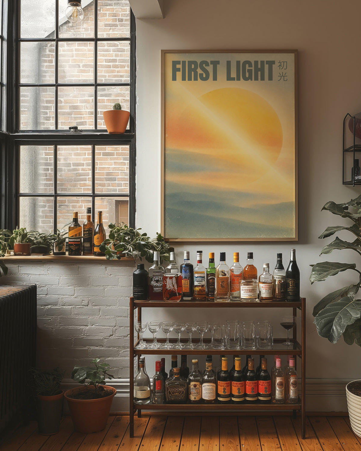First Light Over a Quiet Sea, a pale sunrise over a soft sea, a warm yellow sun glowing above a band of blue water and peach clouds, with the words FIRST LIGHT in gray capitals across the top and a small Japanese word beside them, in a wood frame on a brick wall above a bar cart of bottles and plants