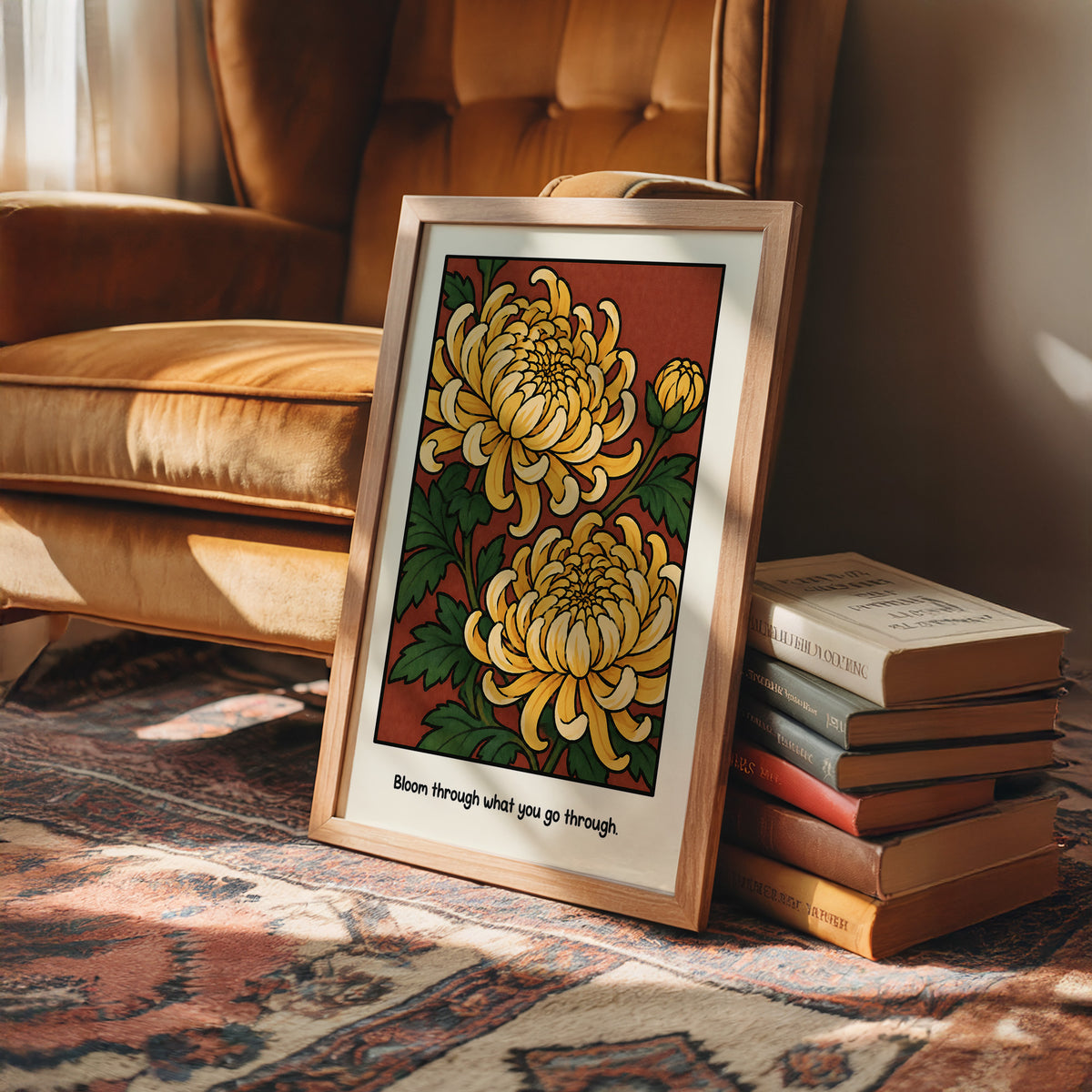 Gold Chrysanthemums Holding Their Ground, a cluster of yellow chrysanthemum blooms with green leaves and a bud on a rust red ground, with the words Bloom through what you go through in small black lettering along the bottom, in a wood frame on a rug beside a leather armchair and a stack of books