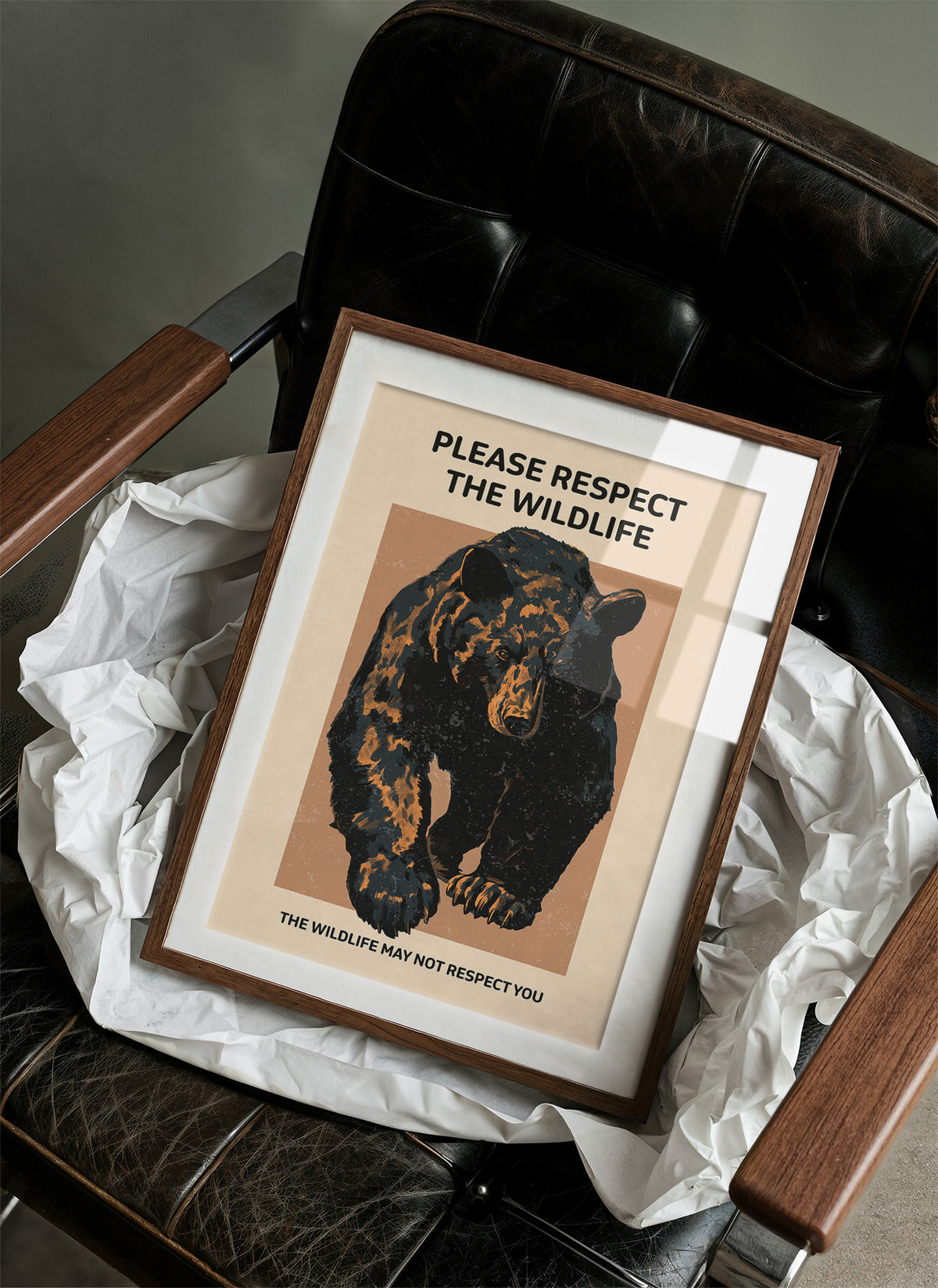 Keep a Respectful Distance, a large black bear with amber and brown fur, head low and shoulders bunched, set on a tan ground under bold black lettering that reads PLEASE RESPECT THE WILDLIFE in a vintage warning sign style, in a wood frame on a black leather armchair beside crumpled white tissue