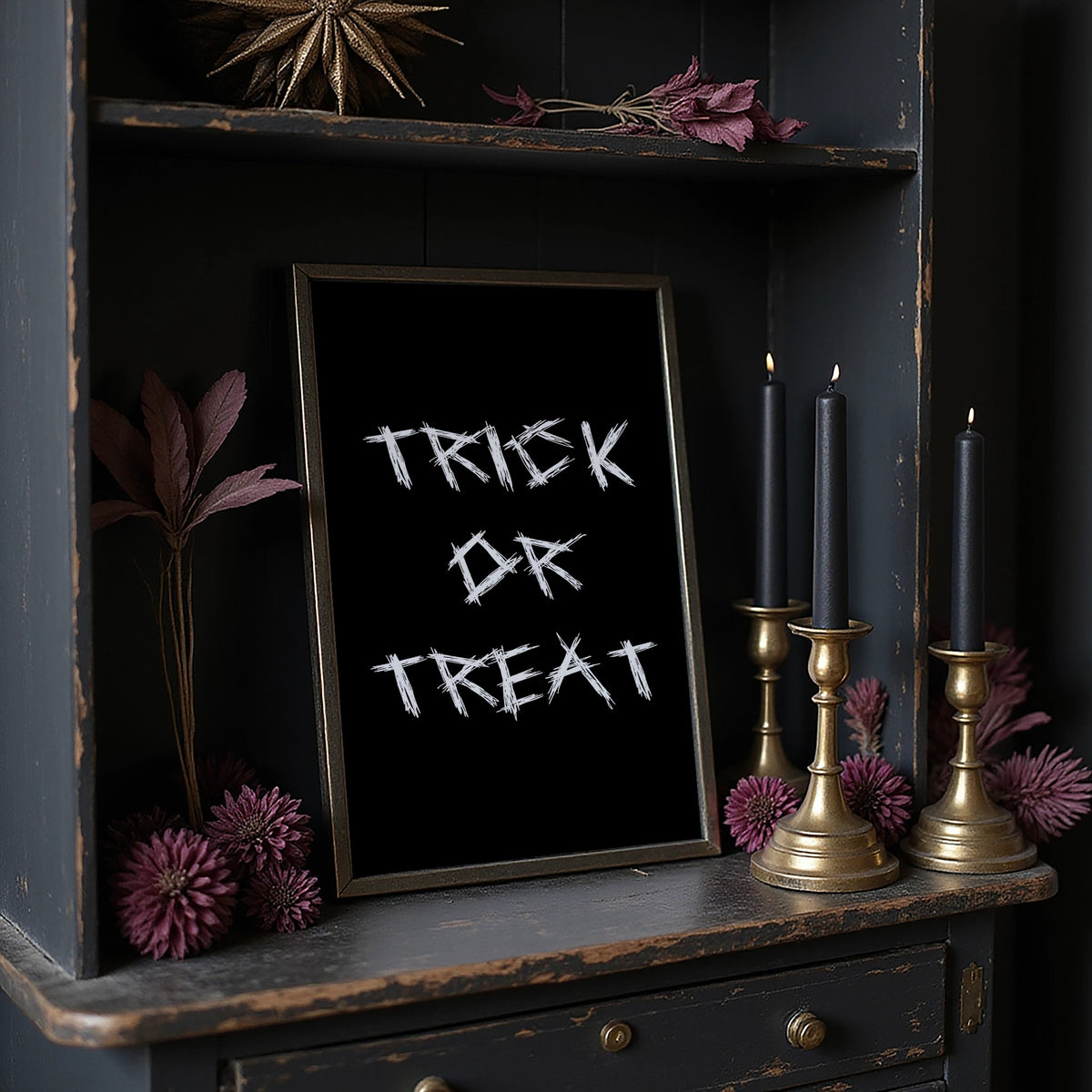 Knock Once for the Sweets, bold chalky white lettering reading TRICK OR TREAT on a solid black ground, hand lettered with a worn brush texture, in a dark frame on a black shelf beside brass candles and dried leaves