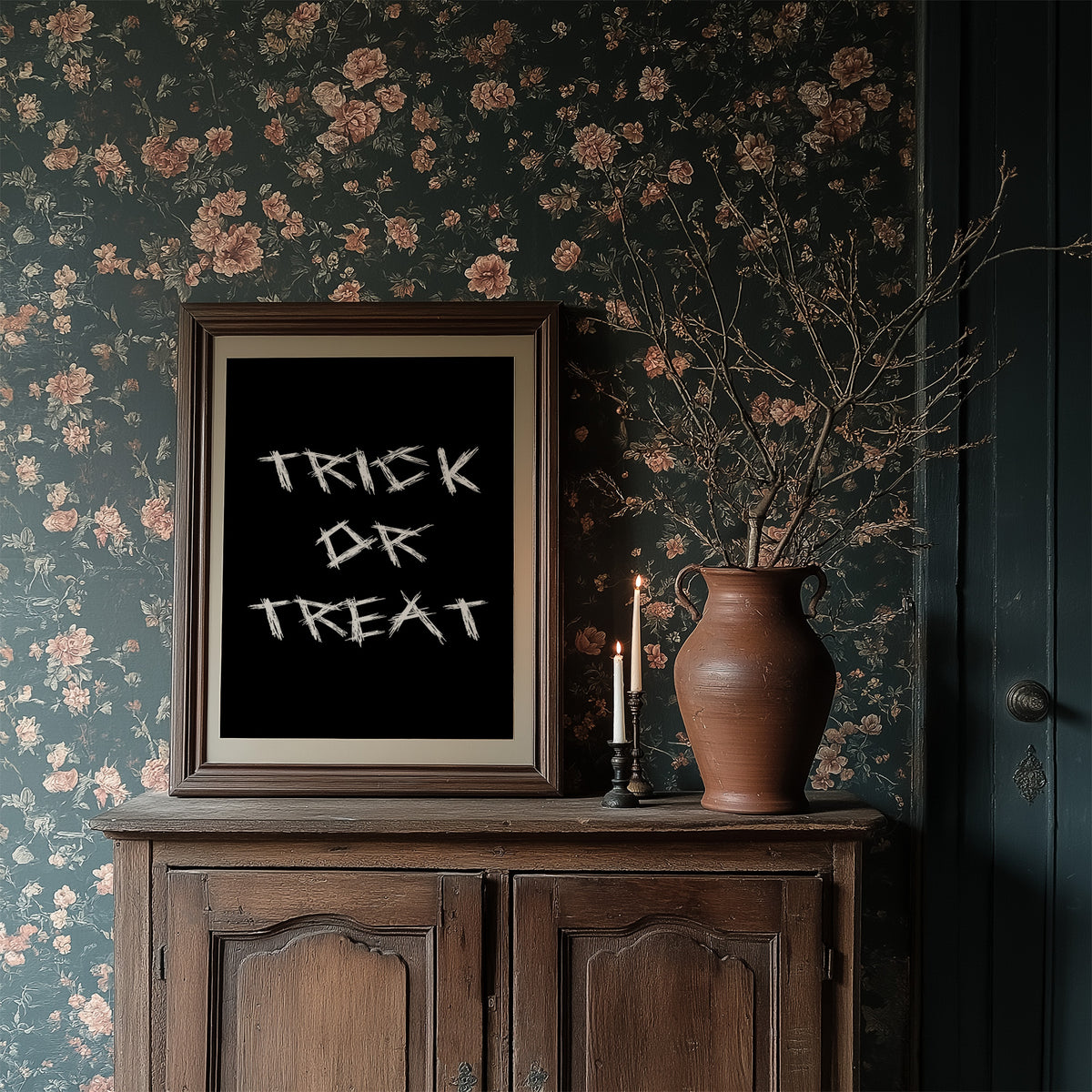 Knock Once for the Sweets, bold chalky white lettering reading TRICK OR TREAT on a solid black ground, hand lettered with a worn brush texture, in a dark wood frame on a floral wallpaper wall above a rustic cabinet and a vase