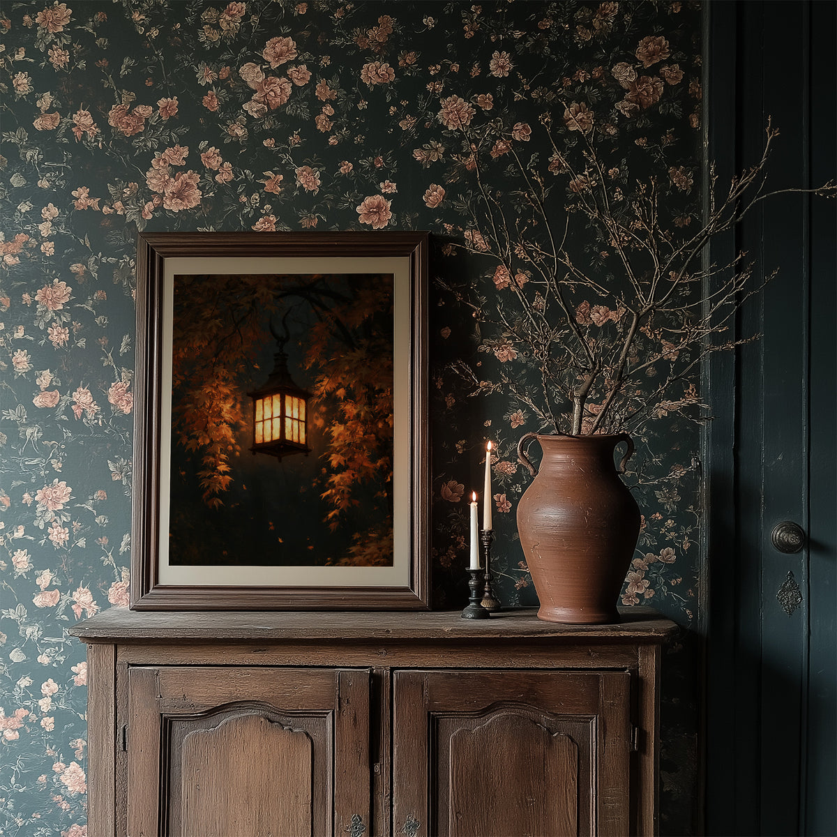 Left On for Someone, a glowing iron lantern hanging in a dark thicket of rust and amber leaves, painted in deep brown, ember orange and warm gold, in a dark wood frame on a floral wallpaper wall above a rustic cabinet and a vase