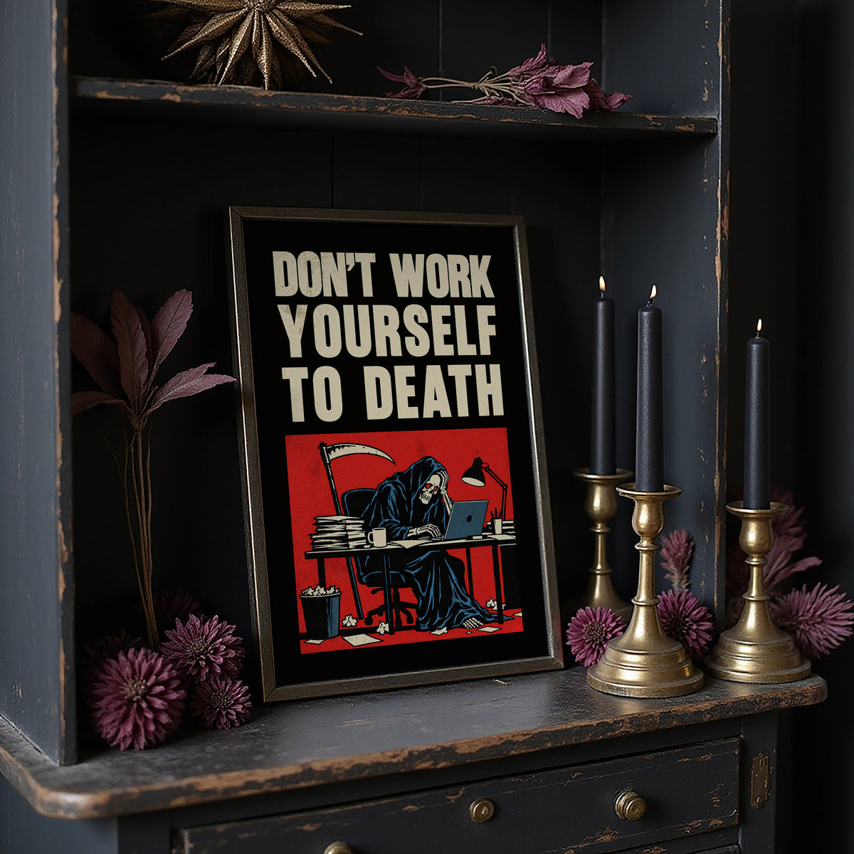 Let the Inbox Wait Till Monday, a hooded reaper hunched over a cluttered desk with a laptop and a stack of files, in a red panel with the lettering DON'T WORK YOURSELF TO DEATH in heavy cream capitals on black, in a dark frame on a black shelf beside brass candles and dried leaves