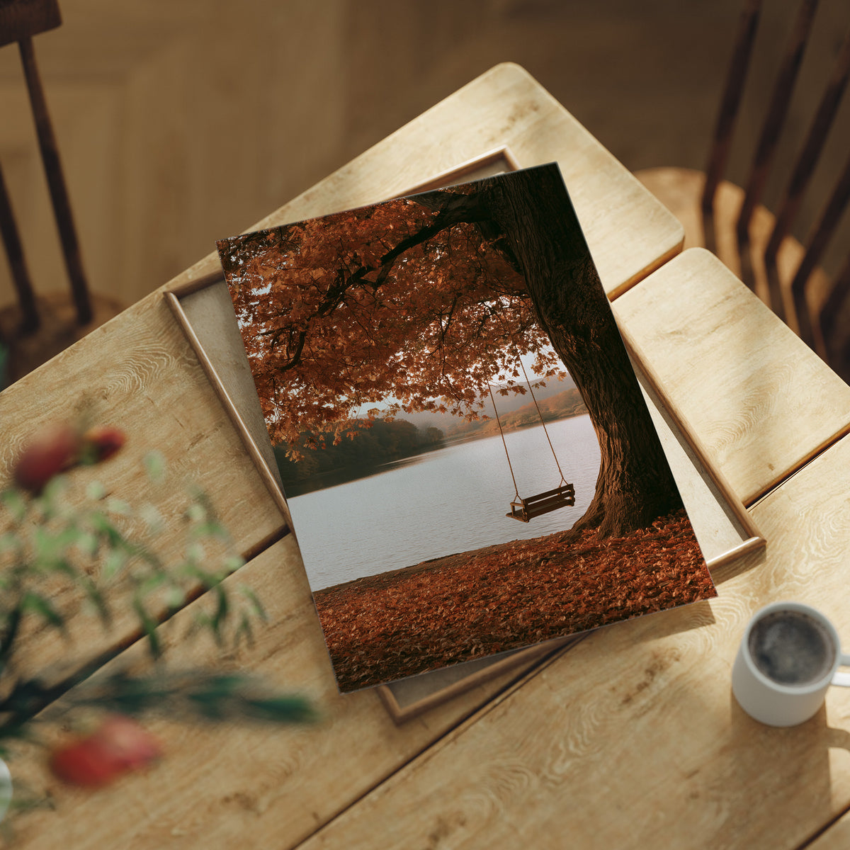 Nobody Has Swung Since Morning, a lone wooden swing hanging from a great tree with orange leaves above a still lake, painted in burnt orange, rust and misty gray, lying flat on a light wood table beside a coffee cup and red berries