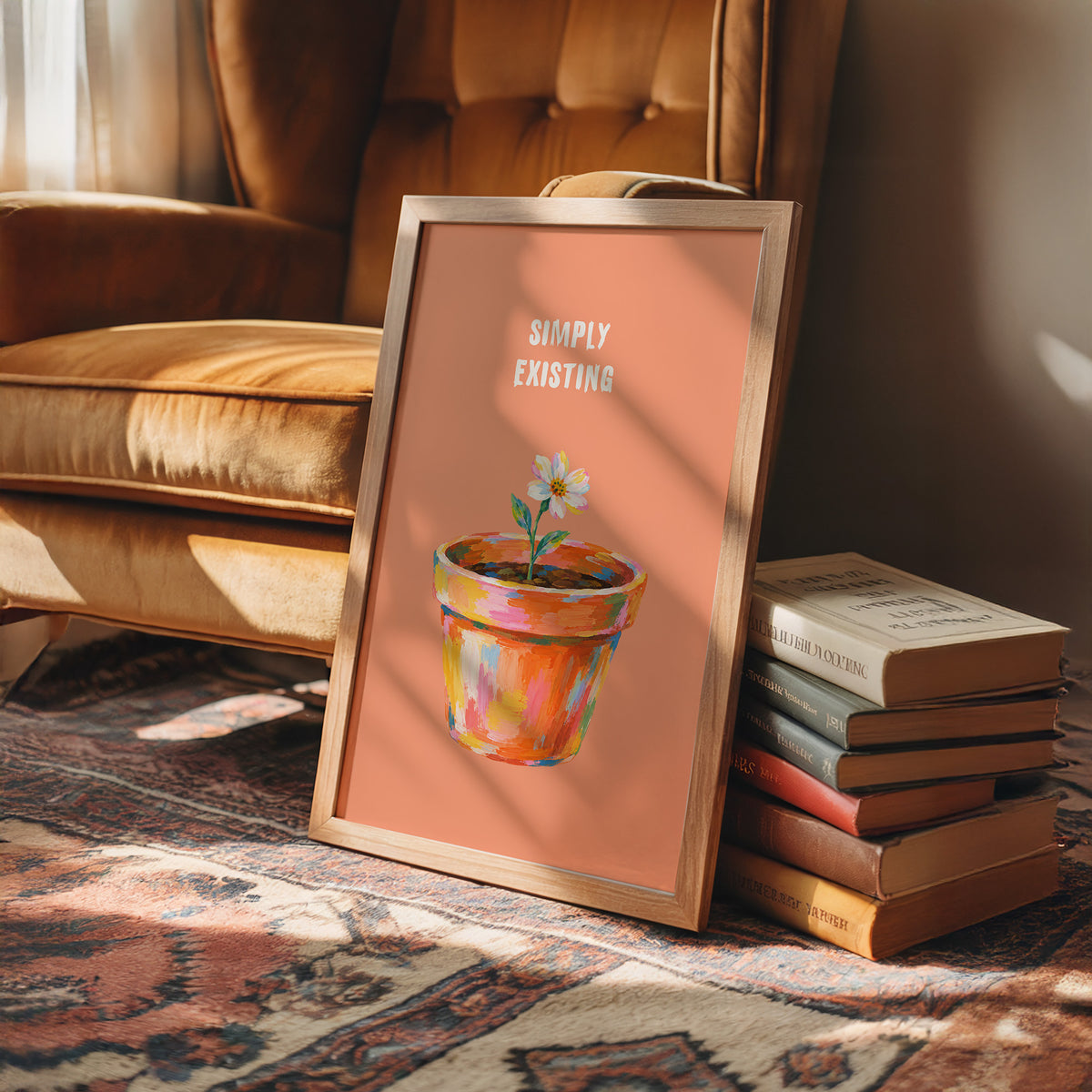 One Small Bloom in Clay, a single white flower rising from a painted clay pot in coral, gold and pink brushstrokes, on a peach ground with two lines of bold pale lettering above, in a loose painterly style, in a wood frame on a rug beside a leather armchair and a stack of books