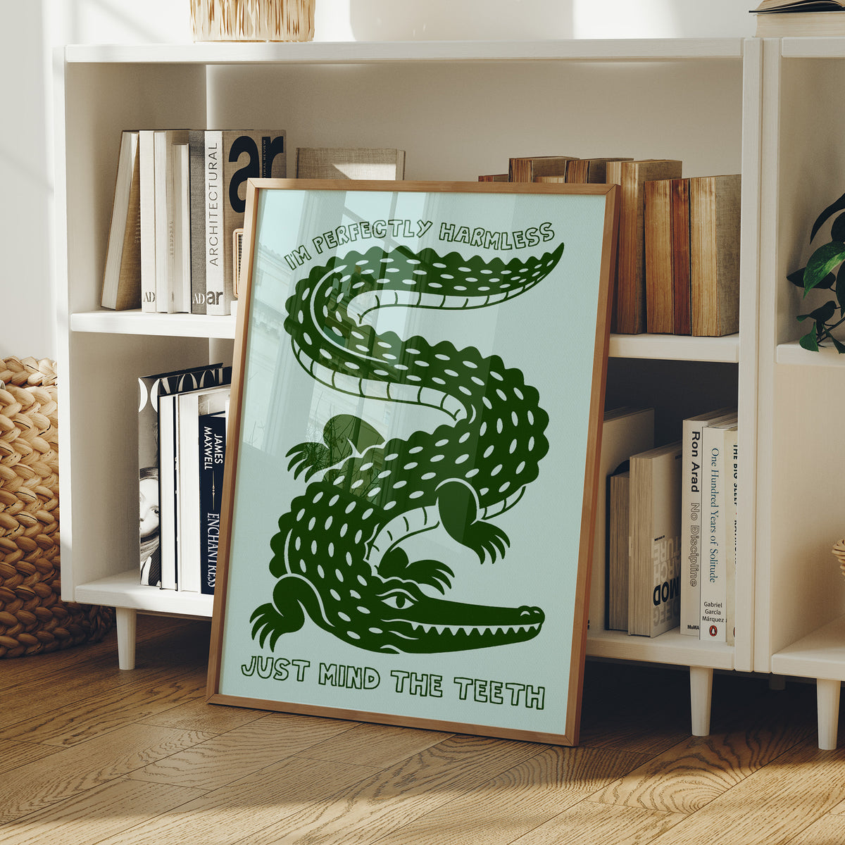 Perfectly Harmless, Mind the Teeth, a deep green crocodile curling down a pale mint ground, with a bold hand lettered line above reading I'M PERFECTLY HARMLESS and one below reading JUST MIND THE TEETH, in a wood frame on a white bookshelf beside stacked books and a small basket
