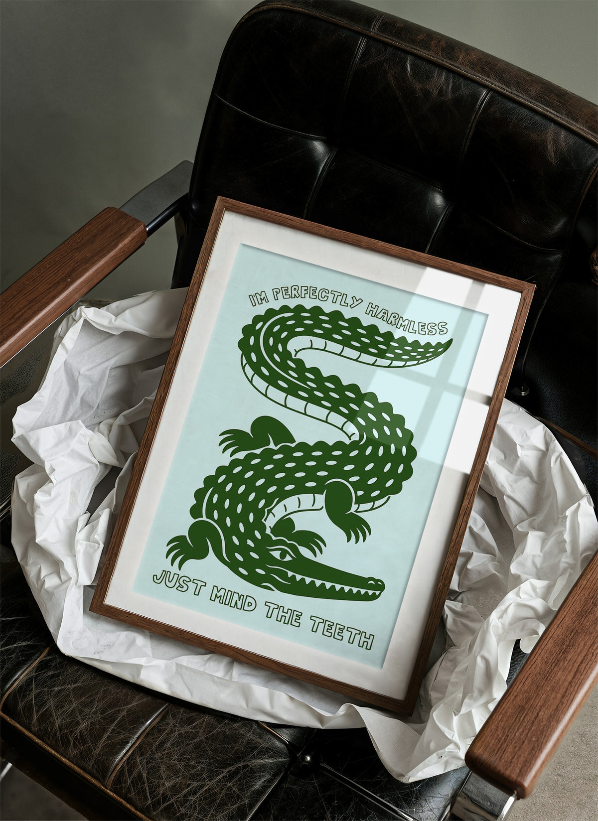 Perfectly Harmless, Mind the Teeth, a deep green crocodile curling down a pale mint ground, with a bold hand lettered line above reading I'M PERFECTLY HARMLESS and one below reading JUST MIND THE TEETH, in a wood frame on a black leather armchair beside crumpled white tissue