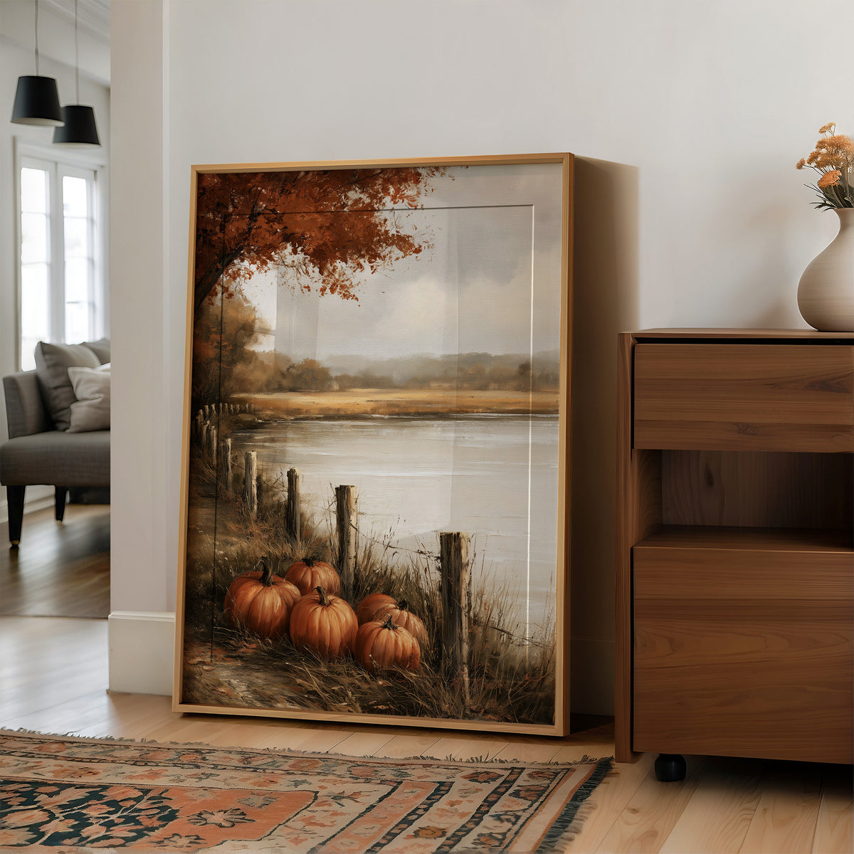 Quiet Water at the Fence Line, weathered fence posts and orange pumpkins on a grassy bank beside a misty lake, with a russet tree leaning over the water, painted in soft ochre, rust and pale gray, in a wood frame on a wall beside a walnut cabinet and an open doorway