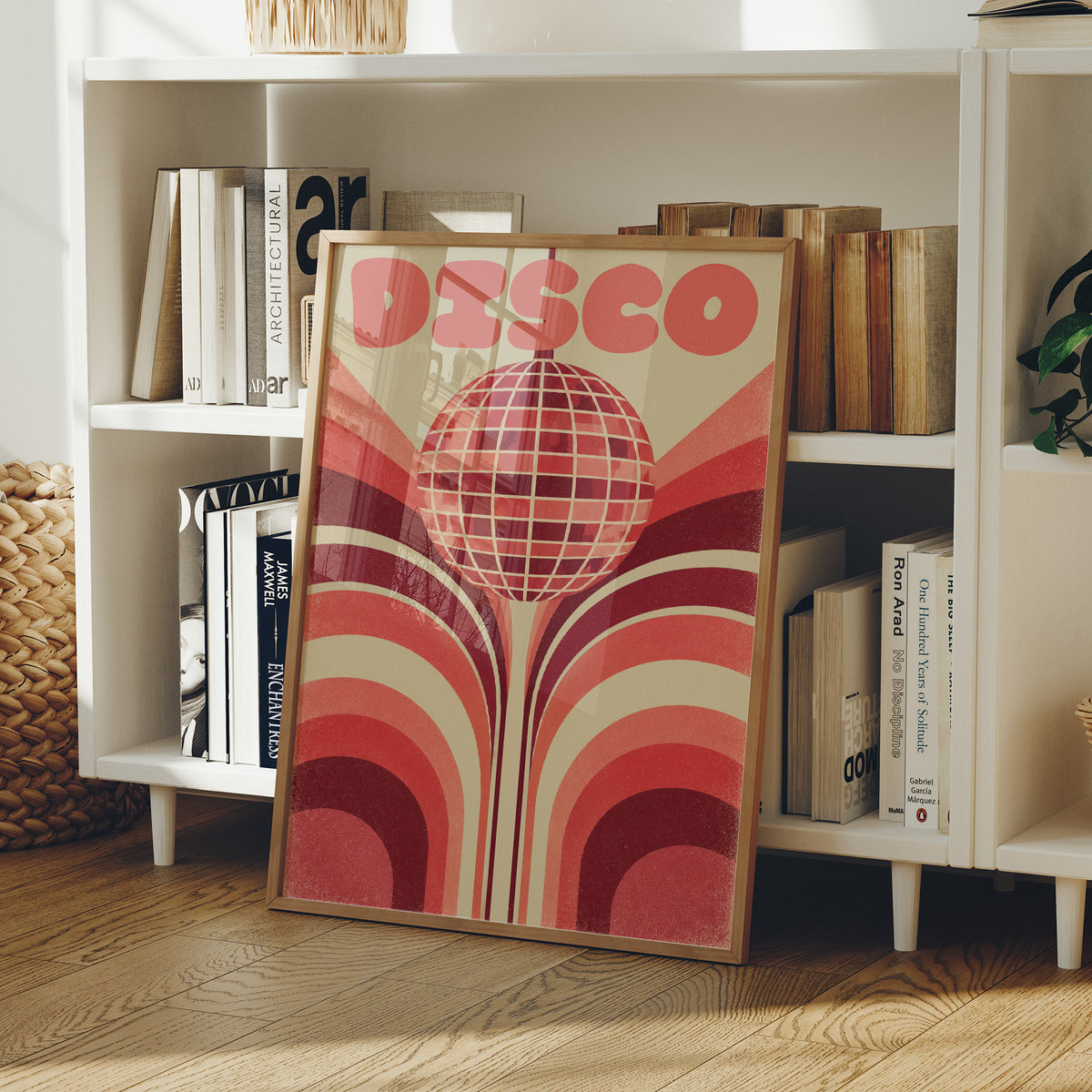 Rose Mirror Ball After Midnight, a retro disco ball drawn in a grid of rose and cream lines at the center of stacked arches in rose, cream and oxblood, with the word DISCO in bold rose letters across the top, in a wood frame on a white bookshelf beside stacked books and a small basket