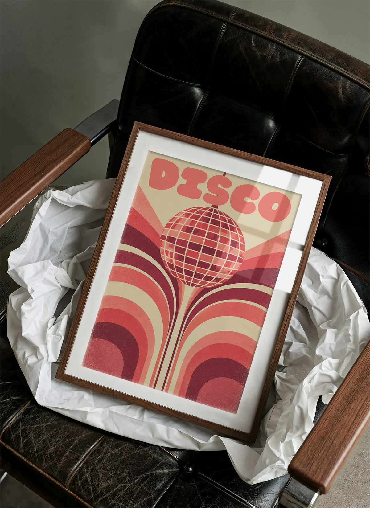 Rose Mirror Ball After Midnight, a retro disco ball drawn in a grid of rose and cream lines at the center of stacked arches in rose, cream and oxblood, with the word DISCO in bold rose letters across the top, in a wood frame on a black leather armchair beside crumpled white tissue