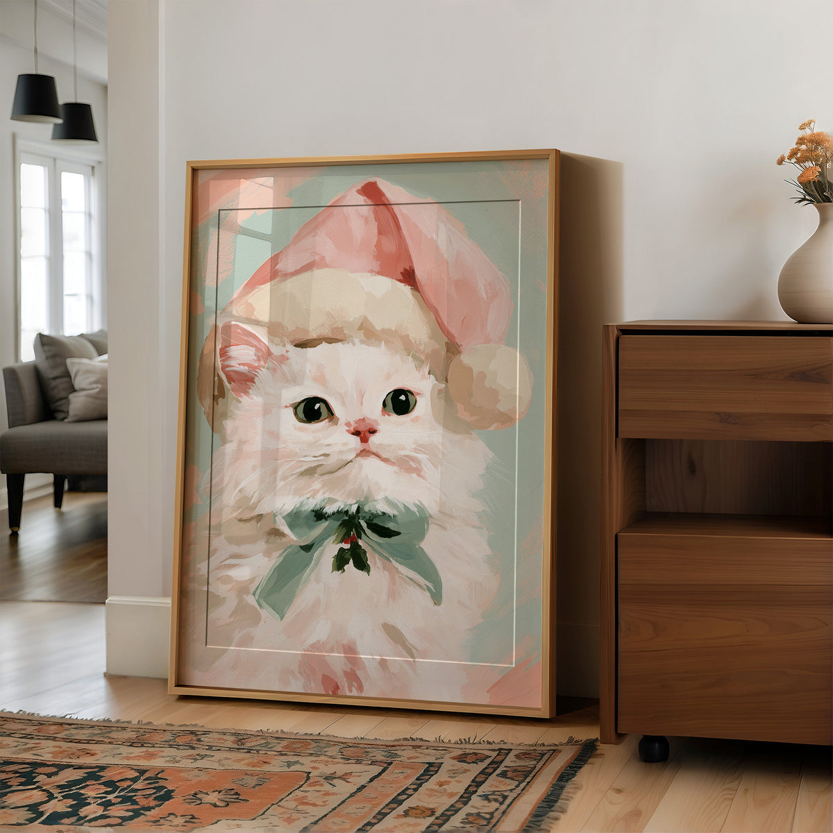 She Dressed Up for the Season, a white kitten in a pink Santa style hat and a mint bow, painted in blush, mint and cream on a soft pastel ground, in a wood frame on a wall beside a walnut cabinet and an open doorway