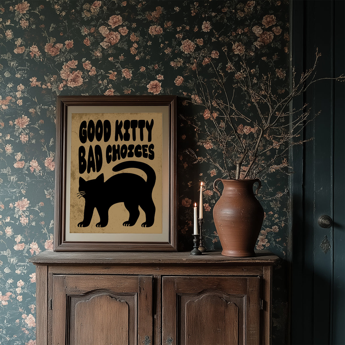 Sleek Silhouette, Questionable Taste, a solid black cat silhouette with a long tail curled up the side, standing on tan paper with the words GOOD KITTY BAD CHOICES in bold black lettering, in a dark wood frame on a floral wallpaper wall above a rustic cabinet and a vase