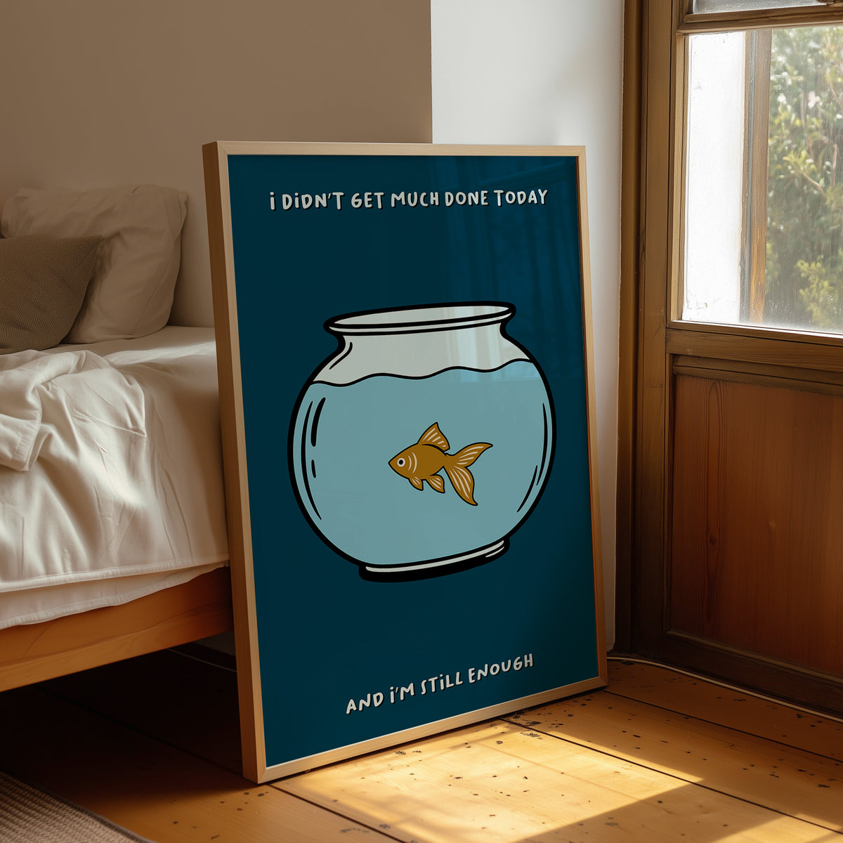 Still Enough for Today, a round glass fishbowl with one goldfish on a deep navy ground, a line of cream lettering above and below in a cartoon style, painted in navy, pale blue and orange, leaning on a wood floor in a bedroom beside white linen bedding