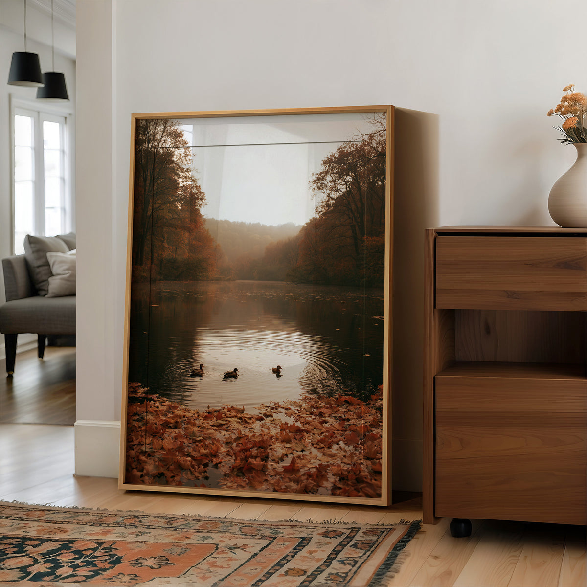 The Pond Keeps Its Quiet Company, two ducks on a still misty lake among rust trees, a bank of fallen autumn leaves in the foreground, painted in rust, forest green and soft gold, in a wood frame on a wall beside a walnut cabinet and an open doorway