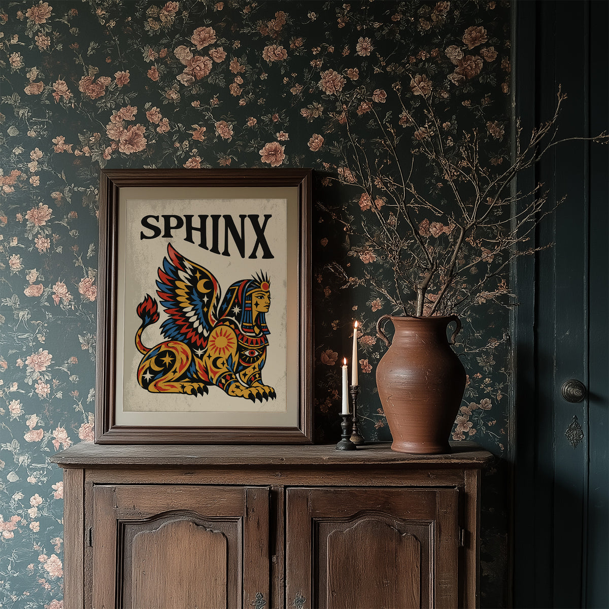 The Sphinx Keeps Her Riddle, a winged sphinx seated with folded wings in gold, red and blue, painted in bold black outlines on a cream paper ground beneath bold black lettering, in a traditional tattoo flash style, in a dark wood frame on a floral wallpaper wall above a rustic cabinet and a vase