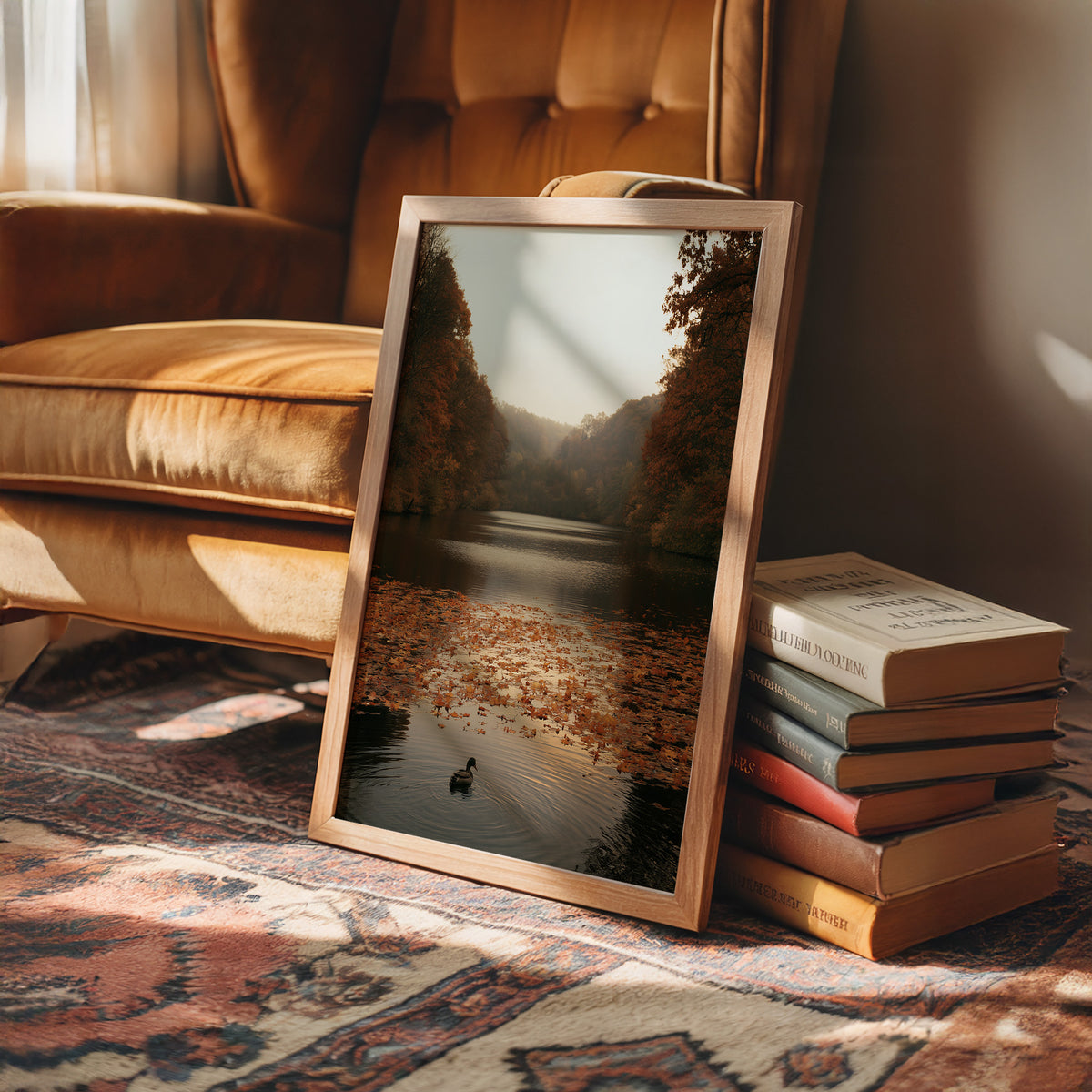 The Swan Waits on the Lake, a lone swan drifting on still water under turning autumn trees, with fallen leaves on the shore and a hazy golden light, painted in rust, amber and soft slate, in a wood frame on a rug beside a leather armchair and a stack of books