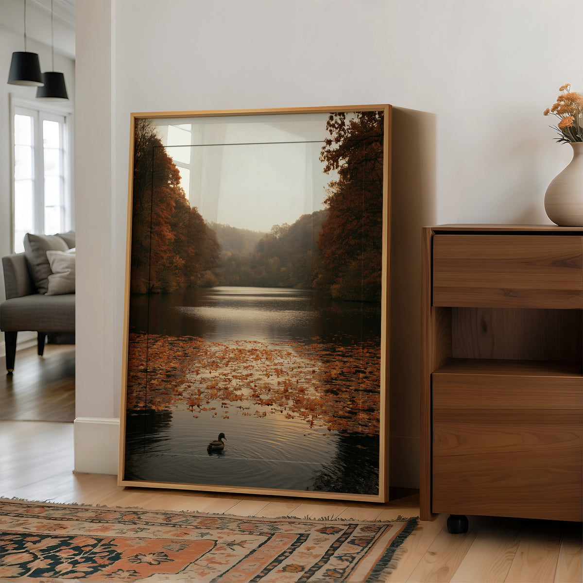 The Swan Waits on the Lake, a lone swan drifting on still water under turning autumn trees, with fallen leaves on the shore and a hazy golden light, painted in rust, amber and soft slate, in a wood frame on a wall beside a walnut cabinet and an open doorway