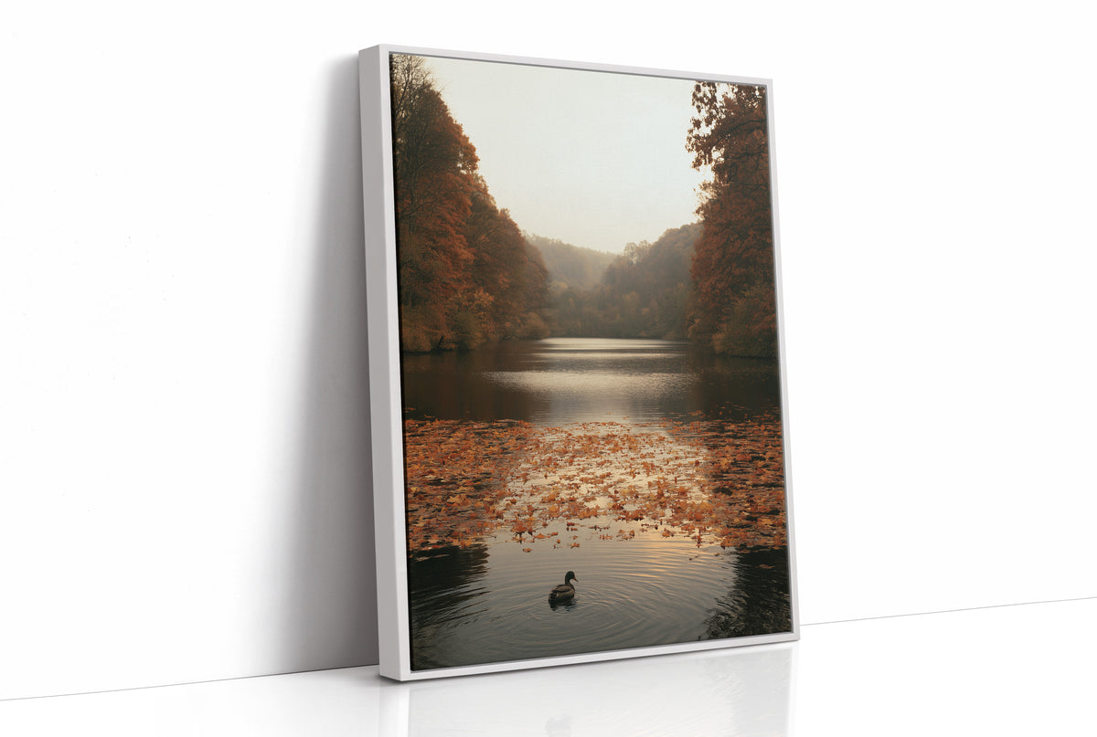 The Swan Waits on the Lake, a lone swan drifting on still water under turning autumn trees, with fallen leaves on the shore and a hazy golden light, painted in rust, amber and soft slate, as a framed canvas in a white frame