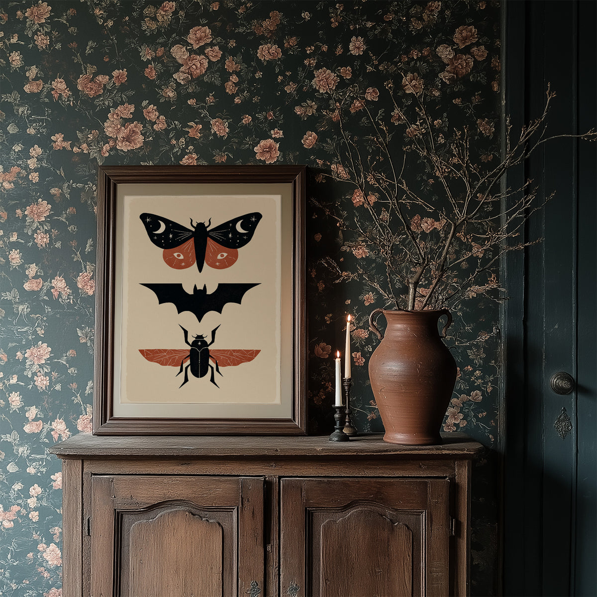 Three Night Visitors in a Row, a moth with crescent moon spots, a black bat in flight and a beetle with rust wings stacked in a column on a cream ground, painted in black, rust and cream, in a dark wood frame on a floral wallpaper wall above a rustic cabinet and a vase