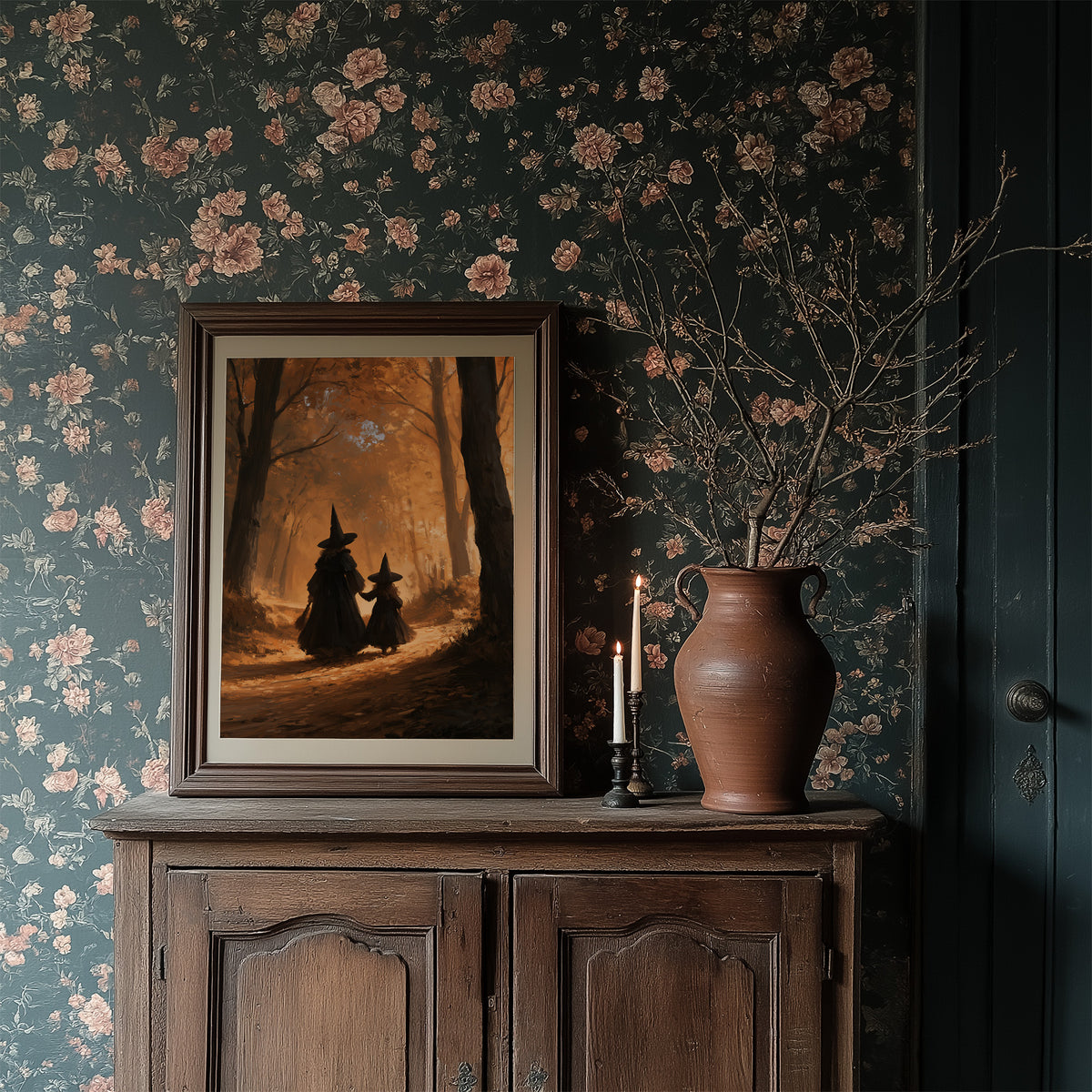 Two Witches on the Amber Path, two witch silhouettes walking a forest path between tall trees, glowing haze and a pale starry cluster above, painted in amber, umber and deep shadow, in a dark frame on a wooden dresser against floral wallpaper
