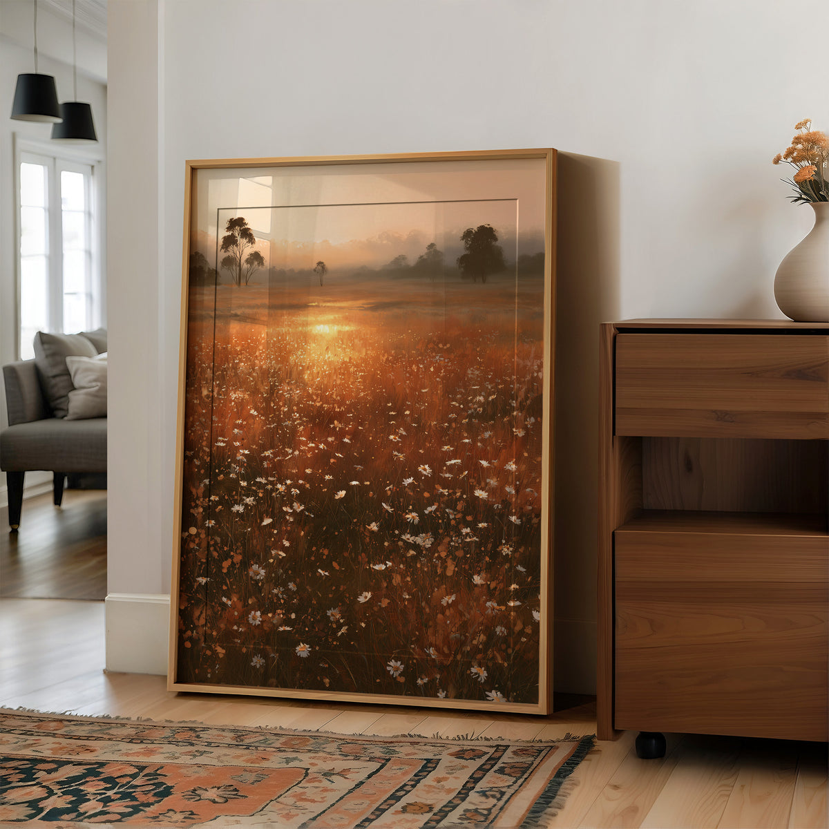 Where the Meadow Holds the Sun, a misty field of wildflowers and grass running toward a low sun, a line of dark trees at the horizon, painted in rust, amber and dusky plum, in a wood frame on a wall beside a walnut cabinet and an open doorway