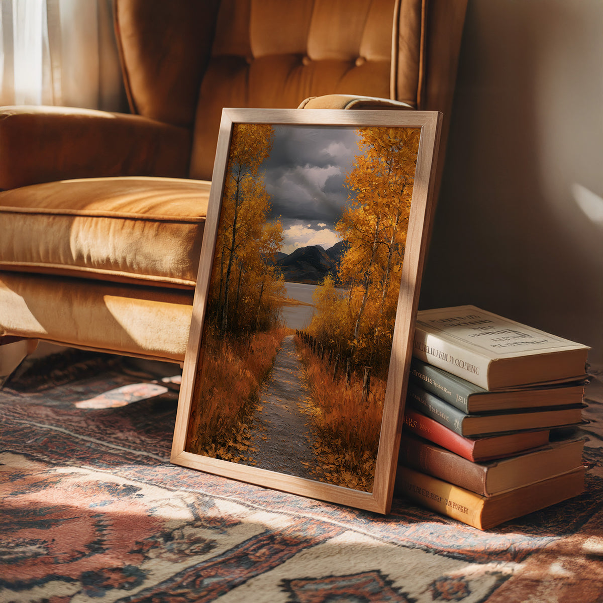Where the Storm Lets Go of the Lane, a dirt lane between gold birches toward soft mountains under a clearing gray sky, painted in amber, slate and rust, in a wood frame on a rug beside a leather armchair and a stack of books