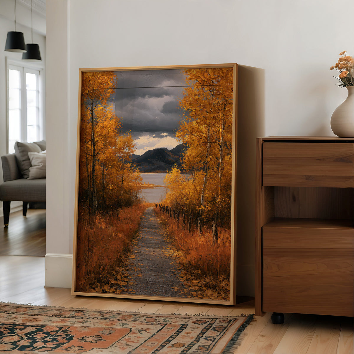 Where the Storm Lets Go of the Lane, a dirt lane between gold birches toward soft mountains under a clearing gray sky, painted in amber, slate and rust, in a wood frame on a wall beside a walnut cabinet and an open doorway
