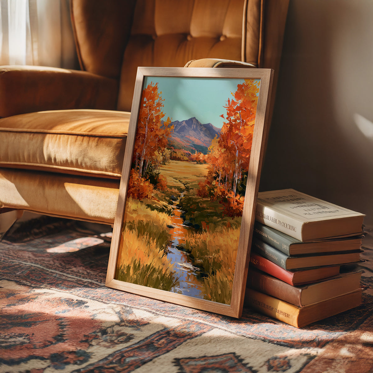 Where the Valley Turns Amber, an autumn valley of aspen trees in amber and rust beside a winding creek, with blue mountains and a pale sky above, painted in amber, rust and sky blue, in a wood frame on a rug beside a leather armchair and a stack of books