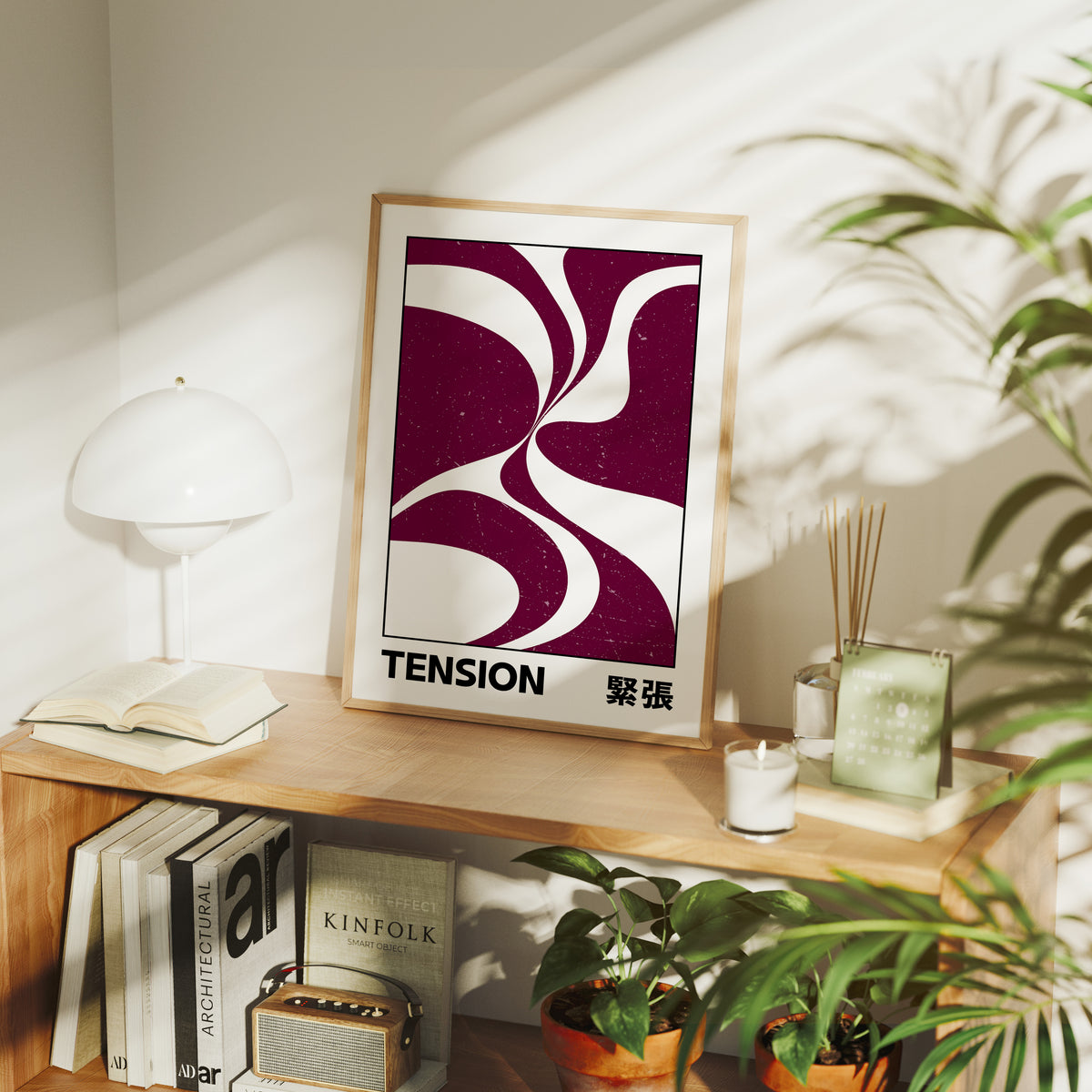 Where Two Curves Almost Touch, two maroon curved forms on a cream ground that lean toward each other and almost meet, with the word TENSION in black capitals and a small Japanese word beside it, in a wood frame on a console table beside a white lamp and stacked books