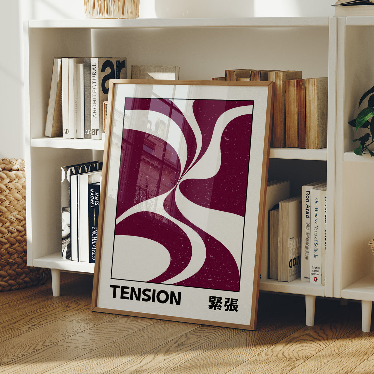 Where Two Curves Almost Touch, two maroon curved forms on a cream ground that lean toward each other and almost meet, with the word TENSION in black capitals and a small Japanese word beside it, in a wood frame on a white bookshelf beside stacked books and a small basket