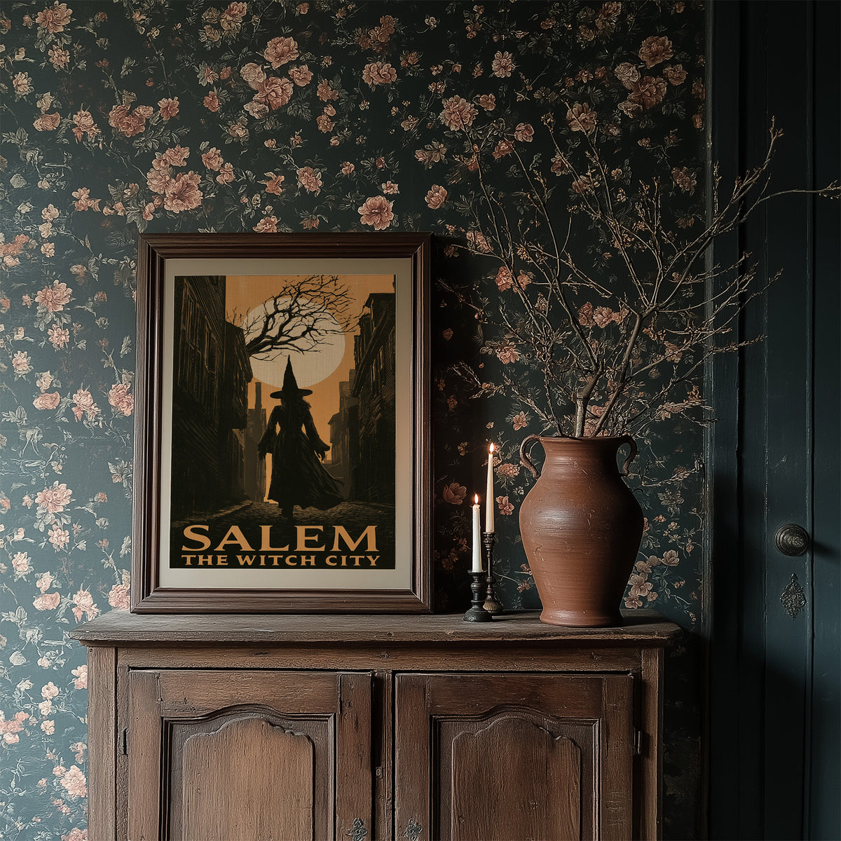 Witch City at Moonrise, a cloaked silhouette beneath a bare tree and a full moon on a vintage style travel poster, painted in amber, olive and black, in a dark frame on a wooden dresser against floral wallpaper