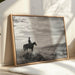 Framed black and white artwork of a person on horseback against a cloudy sky, displayed in a room with natural light.