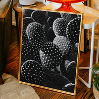 Framed artwork of cacti on a wooden floor with a chair and plants in the background