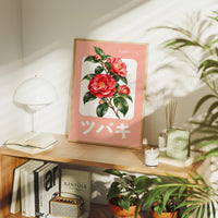 A Camellia Before Noon, a red camellia bloom with green leaves on a blush pink ground, with a rounded cream label and bold pale Japanese lettering, in a flat Japanese travel poster style painted in red, blush pink and cream, in a wood frame on a console table beside a white lamp and stacked books