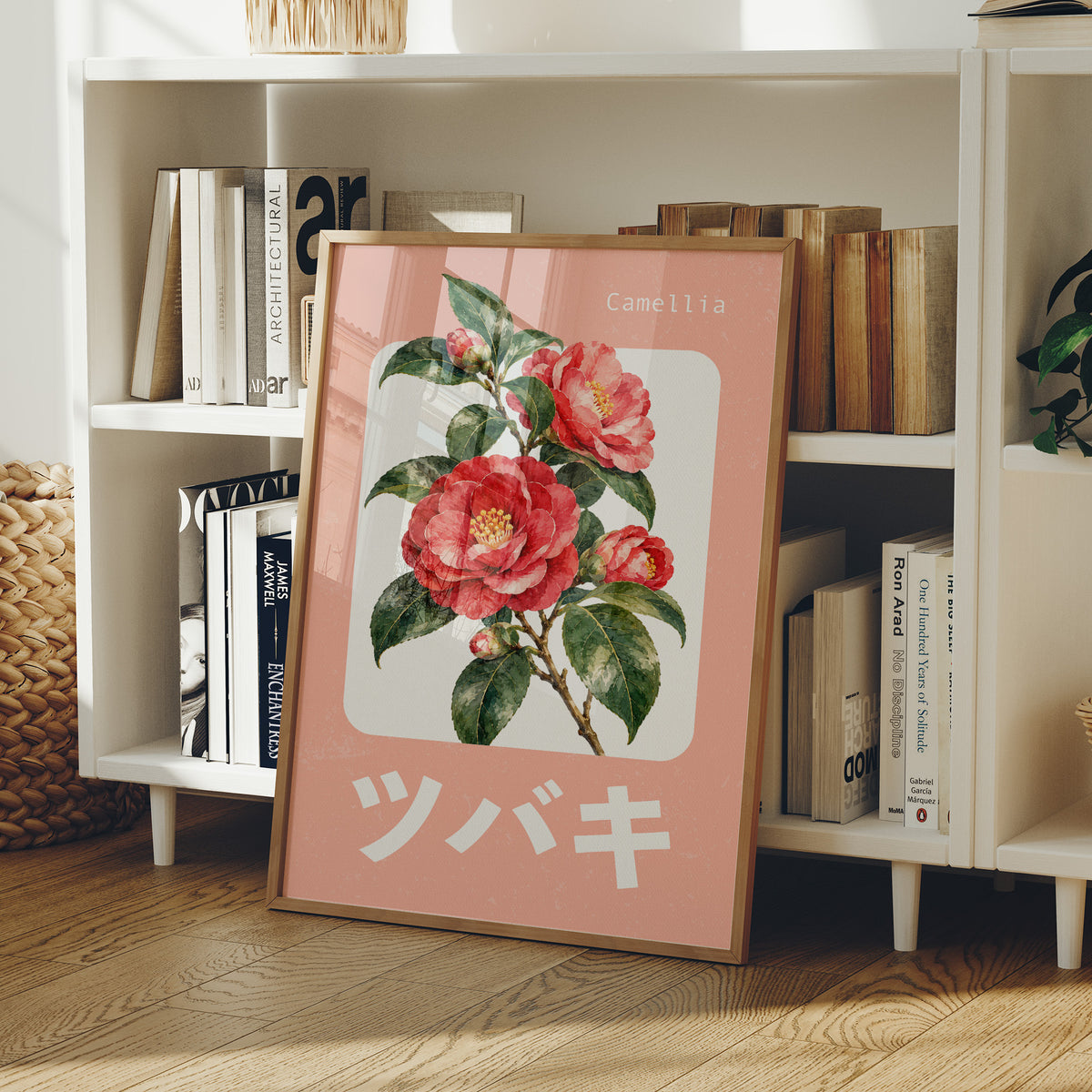A Camellia Before Noon, a red camellia bloom with green leaves on a blush pink ground, with a rounded cream label and bold pale Japanese lettering, in a flat Japanese travel poster style painted in red, blush pink and cream, in a wood frame on a white bookshelf beside stacked books and a small basket
