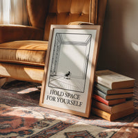 A Room Kept Open for You, a fine black line drawing of a tall empty room with a long floor, a pale window and a small seated figure, with the serif words HOLD SPACE and FOR YOURSELF beneath it on a cream ground, in a wood frame on a rug beside a leather armchair and a stack of books