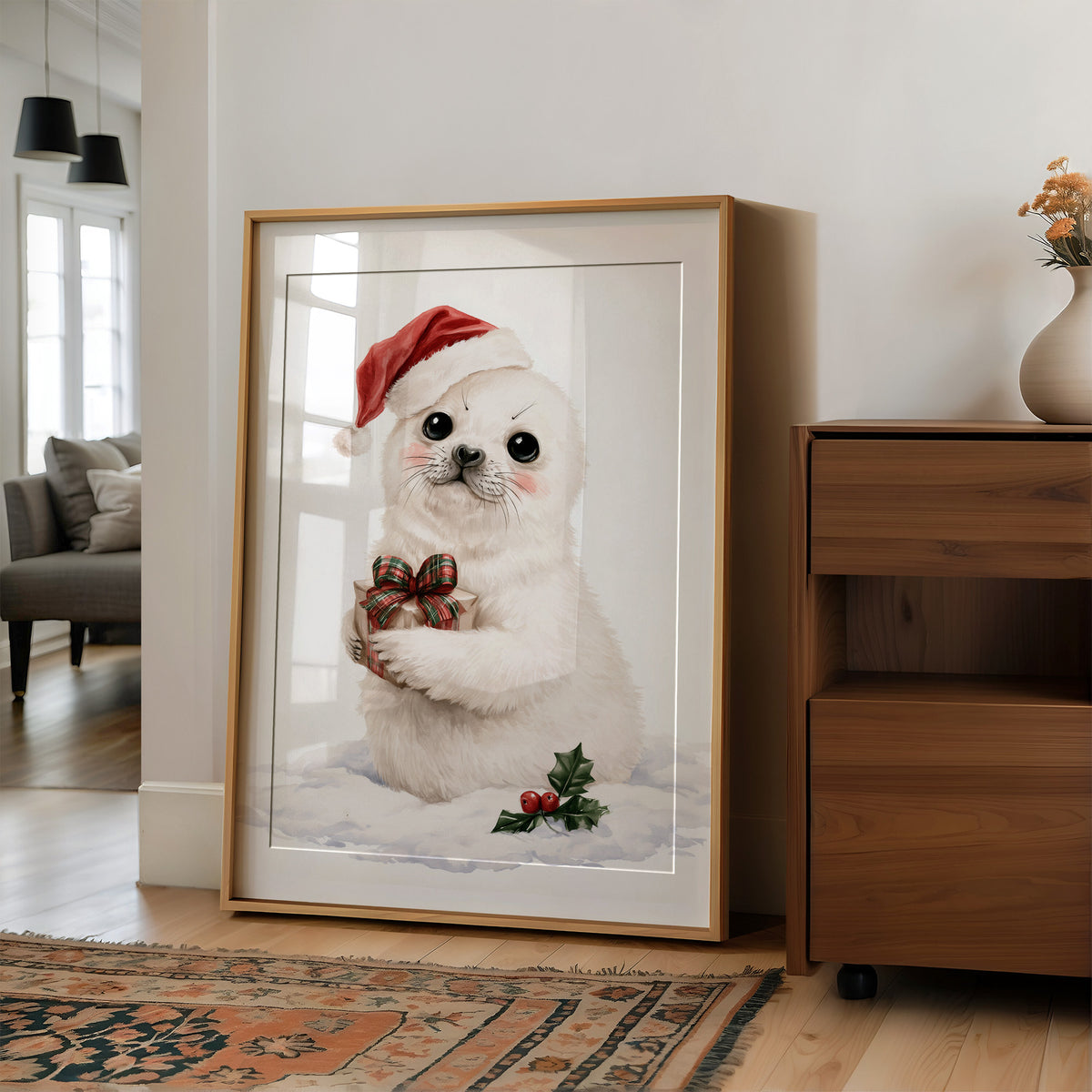 A Snowy Pup Waits for Morning, a white baby seal in a red Santa hat holding a chocolate bow, with a sprig of holly in the snow below, painted in soft cream, red and white, in a wood frame on a wall beside a walnut cabinet and an open doorway