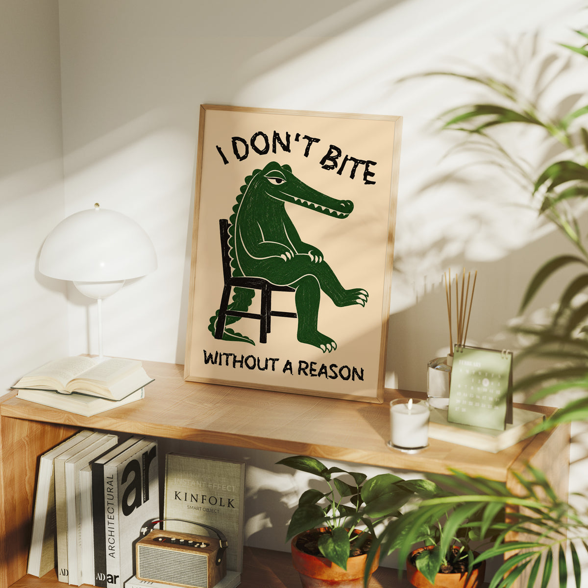 Bites Only With a Reason, a crocodile sitting upright in a plain wooden chair, in deep green on a cream ground, with a bold lettered line above reading I DON'T BITE and one below reading WITHOUT A REASON, in a wood frame on a console table beside a white lamp and stacked books
