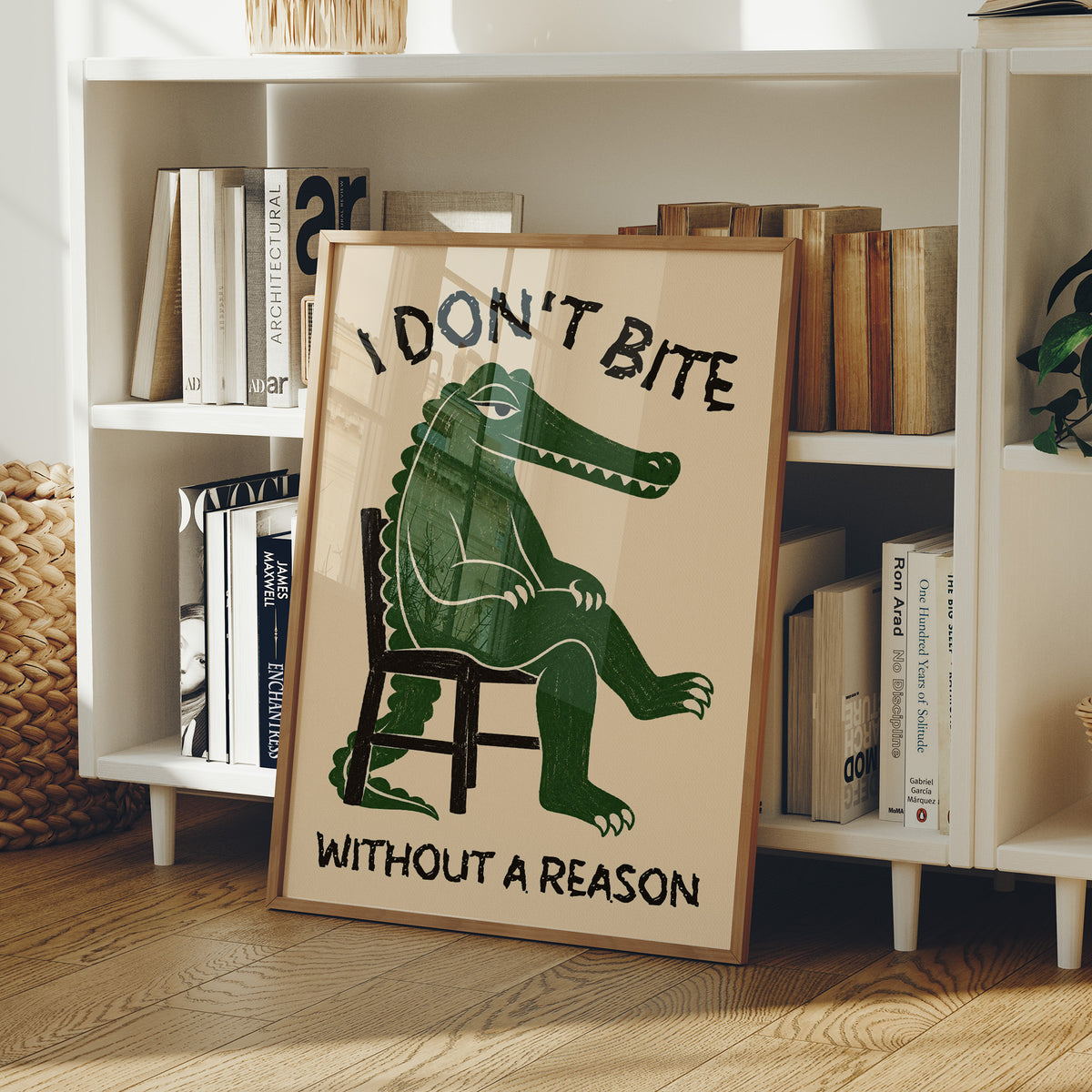 Bites Only With a Reason, a crocodile sitting upright in a plain wooden chair, in deep green on a cream ground, with a bold lettered line above reading I DON'T BITE and one below reading WITHOUT A REASON, in a wood frame on a white bookshelf beside stacked books and a small basket
