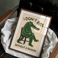 Bites Only With a Reason, a crocodile sitting upright in a plain wooden chair, in deep green on a cream ground, with a bold lettered line above reading I DON'T BITE and one below reading WITHOUT A REASON, in a wood frame on a black leather armchair beside crumpled white tissue