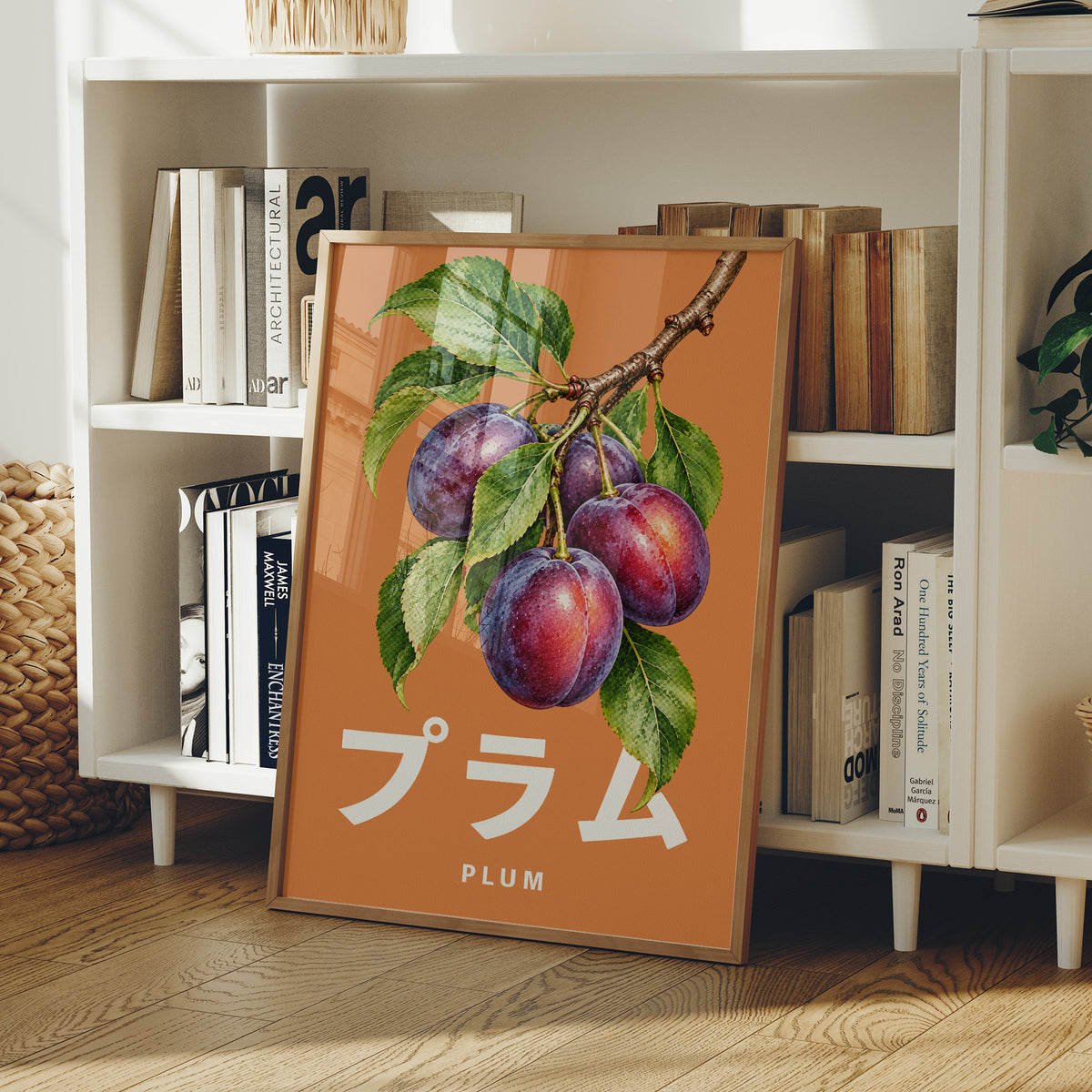 Branch Heavy with Plums, a branch of purple plums with green leaves on a burnt orange ground, with a tall block of Japanese katakana and the English word PLUM set small in cream beneath, in a wood frame on a white bookshelf beside stacked books and a small basket