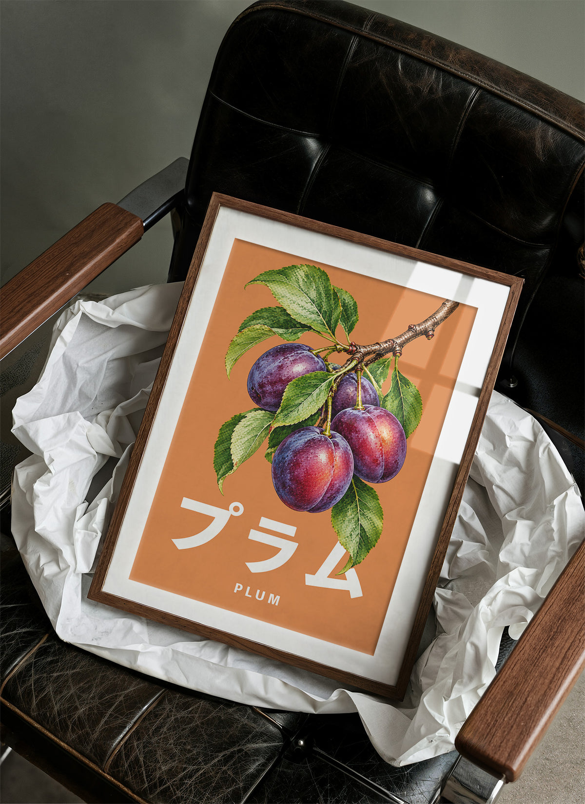 Branch Heavy with Plums, a branch of purple plums with green leaves on a burnt orange ground, with a tall block of Japanese katakana and the English word PLUM set small in cream beneath, in a wood frame on a black leather armchair beside crumpled white tissue